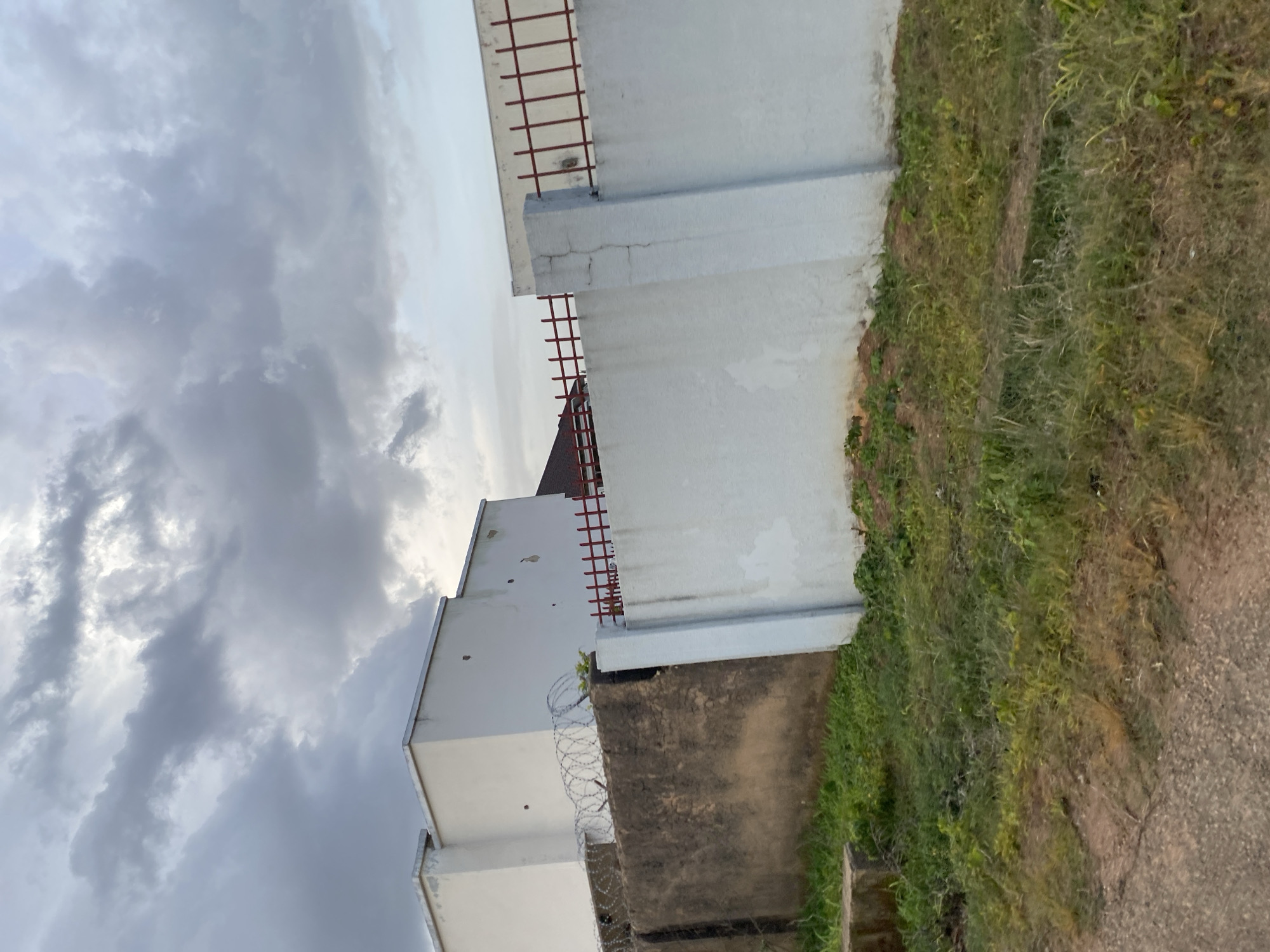 The image depicts a street-level view of a walled property in Barikin Jirga Summa, Nigeria. The walls are constructed from different materials, with a darker, possibly concrete or stucco section on the left, topped with barbed wire. Adjacent to this is a lighter, white-painted wall with a red, barred fence running along the top. Behind the walls, a white building with a flat roof is visible, showing some signs of wear and tear, including dark marks or holes in the facade. The ground in front of the wall is covered with overgrown grass and some bare earth. The sky above is overcast with dramatic, dark clouds, suggesting a potential for rain. The overall impression is of a securely fenced property in an urban or semi-urban environment.
