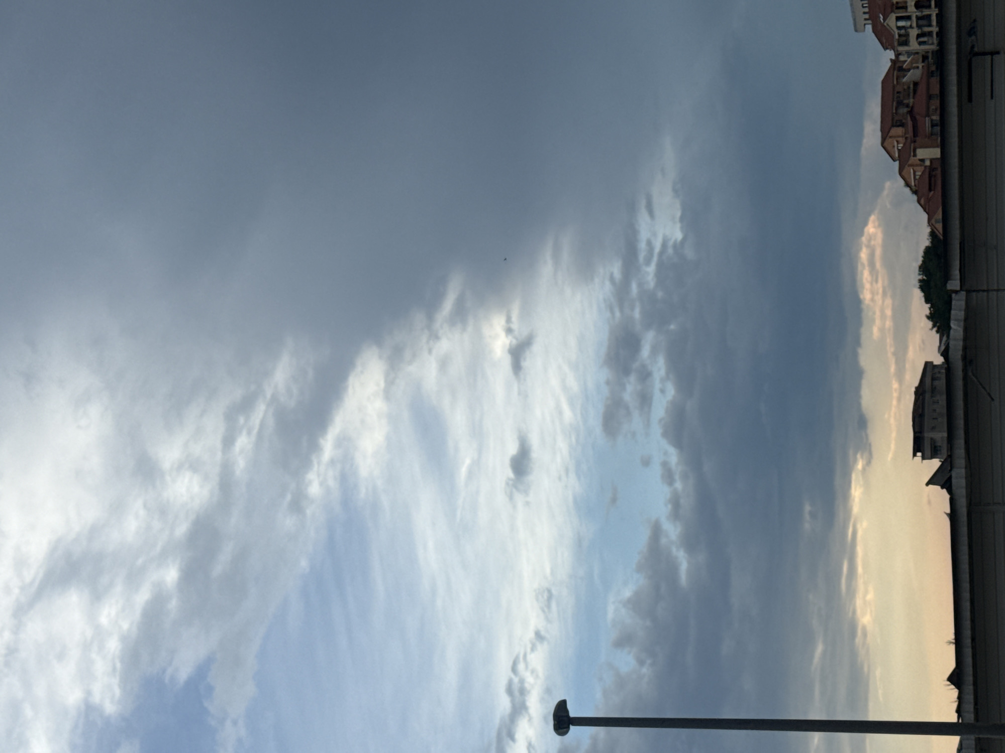 The media file depicts a sky with dramatic cloud formations and a hint of buildings in the upper right corner, suggesting an urban or semi-urban setting. The sky occupies the majority of the frame, with a mix of dark, heavy clouds and lighter, wispy ones. The lighting suggests it is either early morning or late afternoon, possibly nearing sunset, as there's a warm glow in some parts of the sky, contrasting with the cooler, grayer tones of the clouds. A faint silhouette of a bird or insect is visible in the upper portion of the sky. In the upper right, buildings with red tile roofs are visible, and below them, a dark, horizontal line, possibly a roof or structure, spans the frame. A thin, dark pole-like object, also horizontal, is present in the lower portion of the image, potentially a street light or part of a structure. The scene evokes a sense of anticipation, perhaps of an approaching storm or the transition between day and night. No text is visible in the image. The location context provided is Boreti, Montenegro.