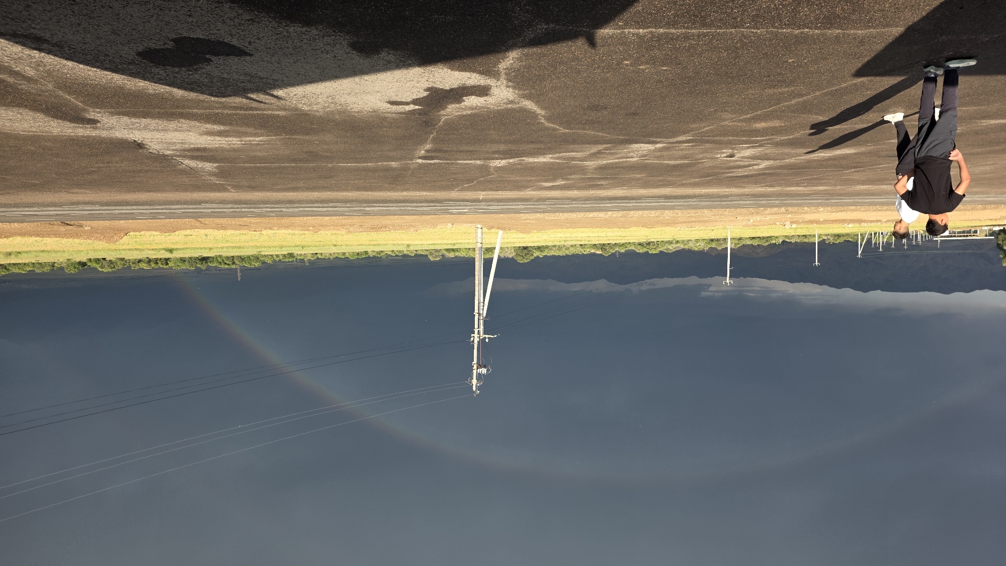 Two young men stand on a cracked asphalt surface, a shadowed area to their right indicating the presence of an unseen object. In the background, a flat, grassy field stretches towards a line of trees and distant, dark mountains under a dramatic, cloudy sky. A faint rainbow arcs across the dark clouds, suggesting recent rainfall. Power lines and poles are visible in the middle ground. The scene appears to be on the outskirts of a city, possibly Bishkek, Kyrgyzstan, given the landscape and power infrastructure.