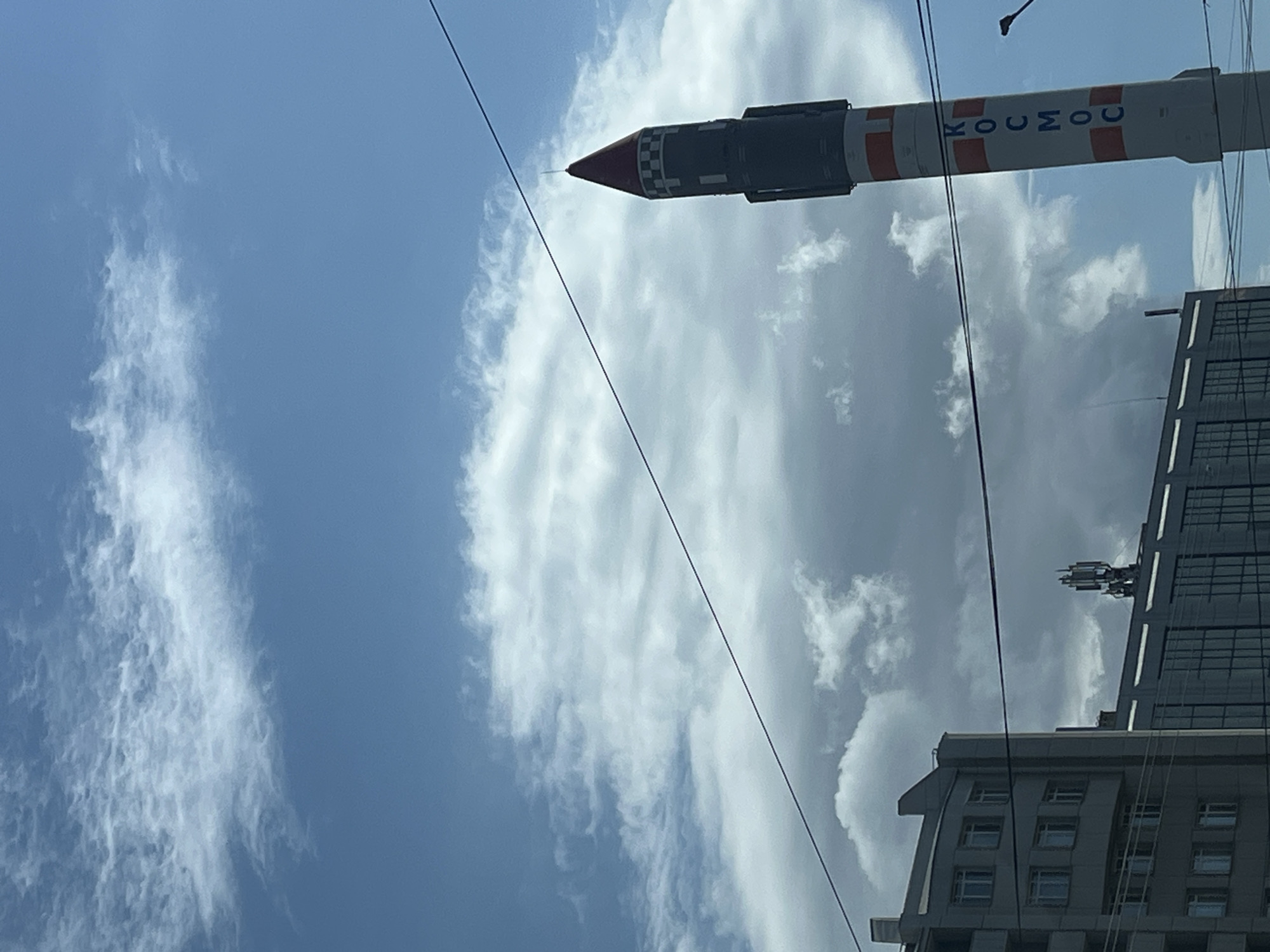 The image shows a partial view of a building and a rocket sculpture against a cloudy blue sky. The rocket is predominantly white with orange stripes and the word "КОСМОС" (Cosmos) written on it. The tip of the rocket is red and black checkered. Electrical wires are visible in the foreground, partially obscuring the view. Part of a tall building with multiple windows is visible on the right side of the image. A communications antenna is attached to the roof of the building.
The image was taken in Krasnoyarsk, Russia.