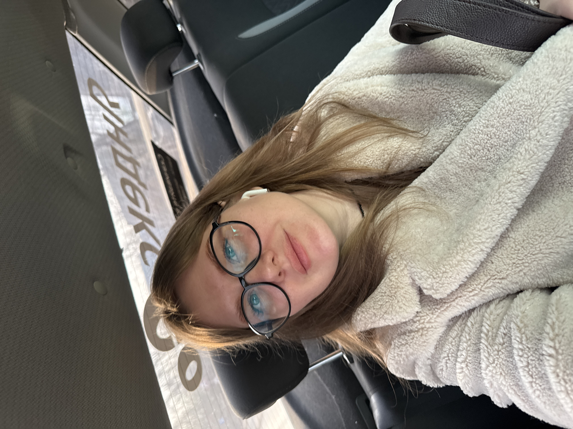 A woman with light brown hair and black-framed glasses is sitting in a vehicle. She is wearing a light-colored, fuzzy jacket. She has an earbud in her left ear. The interior of the vehicle is visible, including the gray leather seats and a headrest. Outside the window, indistinct signage is visible, with the Russian word "Яндекс" (Yandex) partially legible, suggesting the location is Ферма, Russia. A brown leather purse strap is also visible.