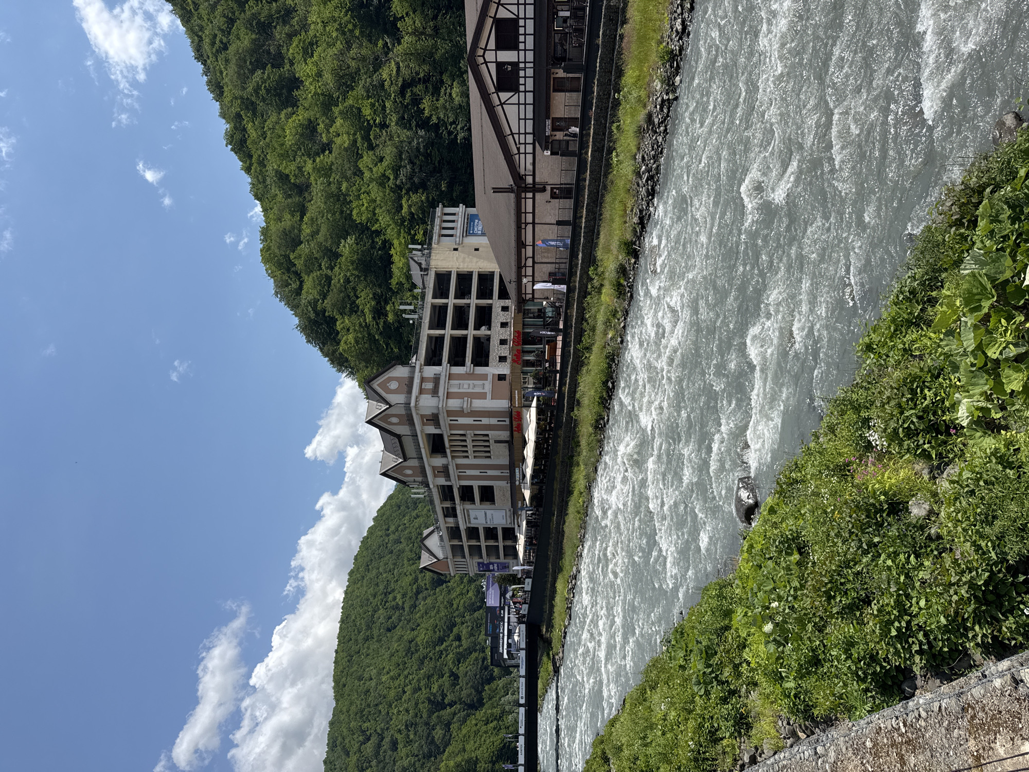 The image shows a wide, fast-flowing river with choppy water, bordered by a grassy embankment on the left and a built-up area on the right. Behind the embankment, a multi-story building with a complex facade, featuring multiple levels of balconies and a distinctive roofline, is visible. Adjacent to this building is a lower structure with a darker roof, appearing to be a commercial establishment with outdoor seating. Further to the left, a bridge spans the river, with traffic and pedestrian activity suggested on it. The background is dominated by lush green, tree-covered mountains under a bright blue sky with scattered white clouds. The scene depicts a developed area in a mountainous, natural environment, likely a resort or town. The location is identified as Estosadok, Russia. There are no discernible individuals in the image, but the presence of the buildings and bridge suggests human activity and infrastructure. The overall impression is of a vibrant day in a scenic location.