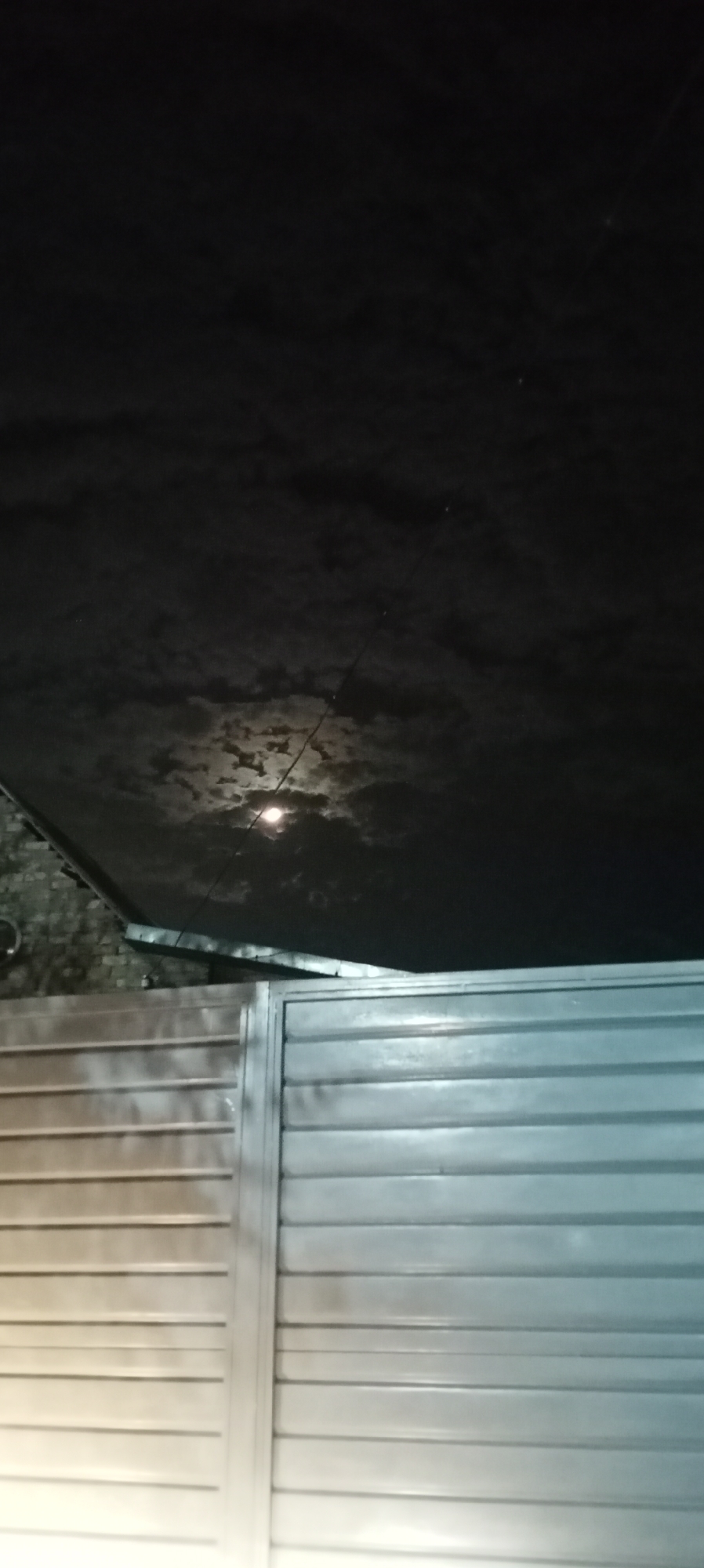 The image depicts a nocturnal scene in Sokuluk, Kyrgyzstan. A metallic gate, likely part of a residential or commercial property, occupies the lower half of the frame. The gate is composed of horizontal slats and appears to be either a sliding or hinged door, reflecting ambient light. Above the gate, the sky is visible, partially obscured by dark, textured clouds. A bright, luminous orb, possibly the moon, is partially visible through a break in the clouds, casting a soft glow. A thin, dark line, likely a wire or cable, stretches across the sky. The overall atmosphere is dark and atmospheric, typical of a nighttime landscape.