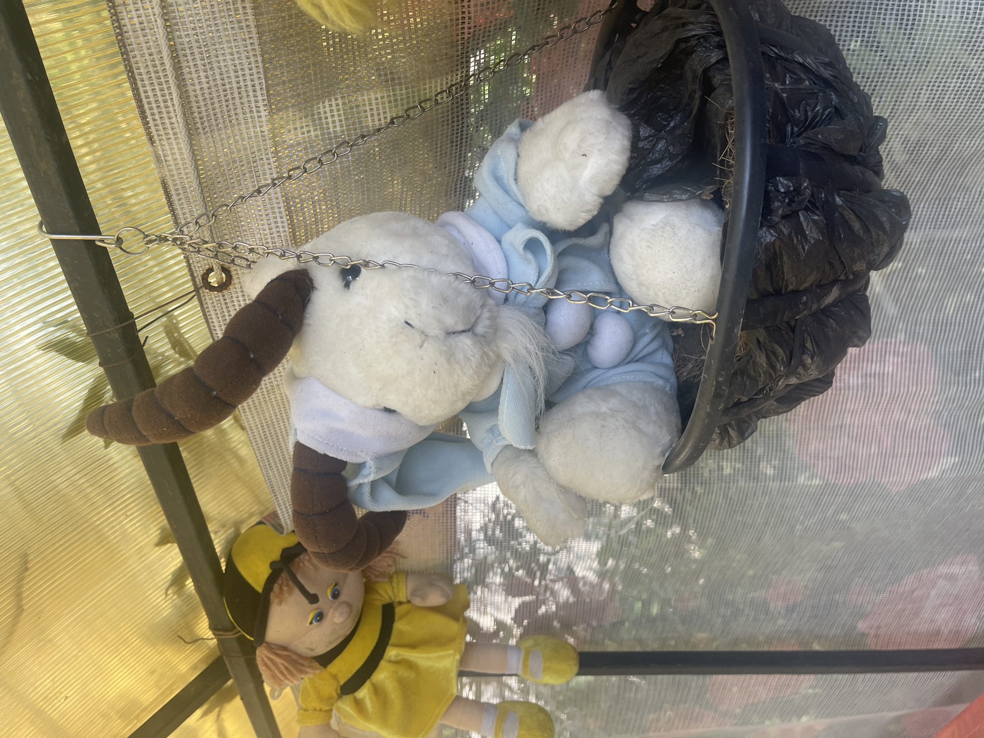 The image shows two stuffed toys hanging inside what appears to be a greenhouse or similar structure. On the left, a doll dressed as a bee, with a yellow dress and black stripes, hangs from a thin wire. Its hair is reddish-brown and its eyes are blue. To the right of the bee doll, a white stuffed goat wearing a light blue outfit is seated in a black hanging basket. The goat has brown, curved horns and a white beard. The basket is lined with black plastic and some dried plant material. The structure has a yellow, translucent roof with visible corrugated texture. A metal frame runs horizontally above the toys, from which they are suspended. The background is a blurred view of foliage and possibly flowers, suggesting an outdoor or garden setting. The image is taken from a slightly low angle, looking up at the toys. The overall scene is domestic and appears to be in an outdoor setting, possibly a garden shed or greenhouse in the city of Sibirskiy, Russia.