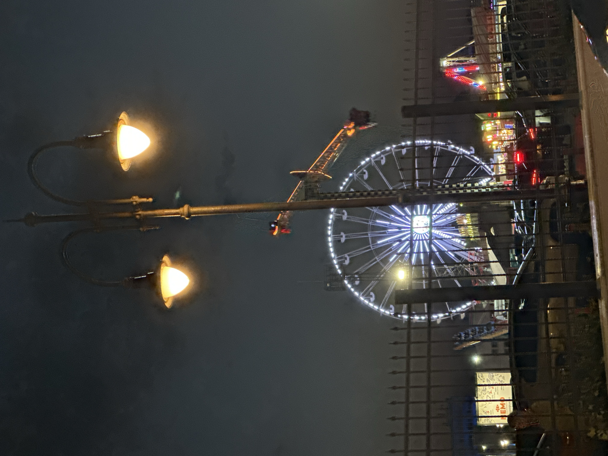A lamppost with two illuminated bulbs stands in the foreground against a dark, cloudy night sky. Behind it, a Ferris wheel with white and blue lights is visible. To the right of the Ferris wheel, a carnival ride with red and yellow lights is in motion, with a passenger visible in one of the gondolas. A black metal fence runs horizontally across the lower portion of the image, obscuring some of the foreground. In the background, several cars are parked, and the lights of other carnival attractions are visible. The scene appears to be a nighttime view of a carnival or amusement park.