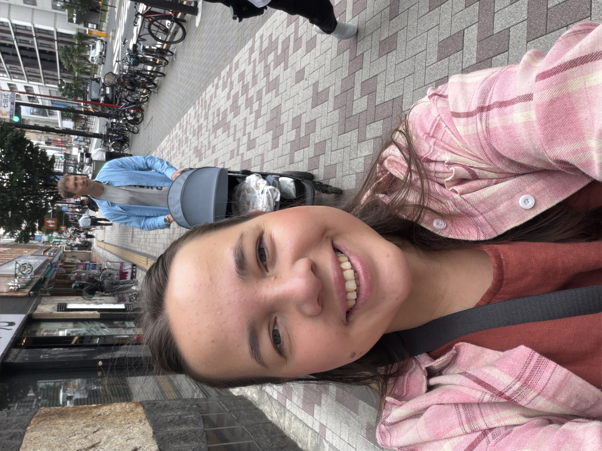 In a city street scene in Meguro, Japan, a woman with dark hair is taking a selfie in the foreground. She is wearing a pink plaid shirt over an orange top and has a grey strap across her shoulder. Behind her, a man in a blue button-up shirt and grey t-shirt is pushing a light grey stroller. The stroller contains bedding. The sidewalk is paved with a grey and pink geometric pattern. To the right of the sidewalk, a row of parked bicycles is visible. Buildings line the street, with storefronts and windows. In the background, cars and pedestrians are present on the street and sidewalk. The sky is overcast.