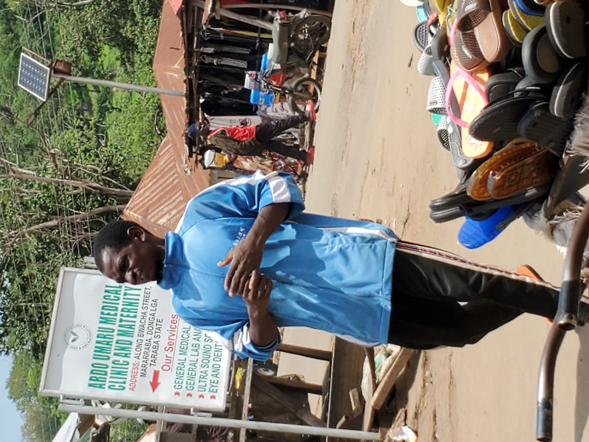 The image shows a street scene in Mararaba, Nigeria, featuring a man standing in front of a sign for "ARDO UMARU MEDICAL CLINIC AND MATERNITY," which advertises general medical, lab, ultrasound, and eye/dental services. The sign indicates the clinic's address is on Bwacha Street in the Donga Local Government Area of Taraba State. The man is wearing a blue jacket and black pants. Behind him, a motorcycle is parked, and a vendor's stall displays an assortment of sandals. A solar panel on a pole is visible in the background.