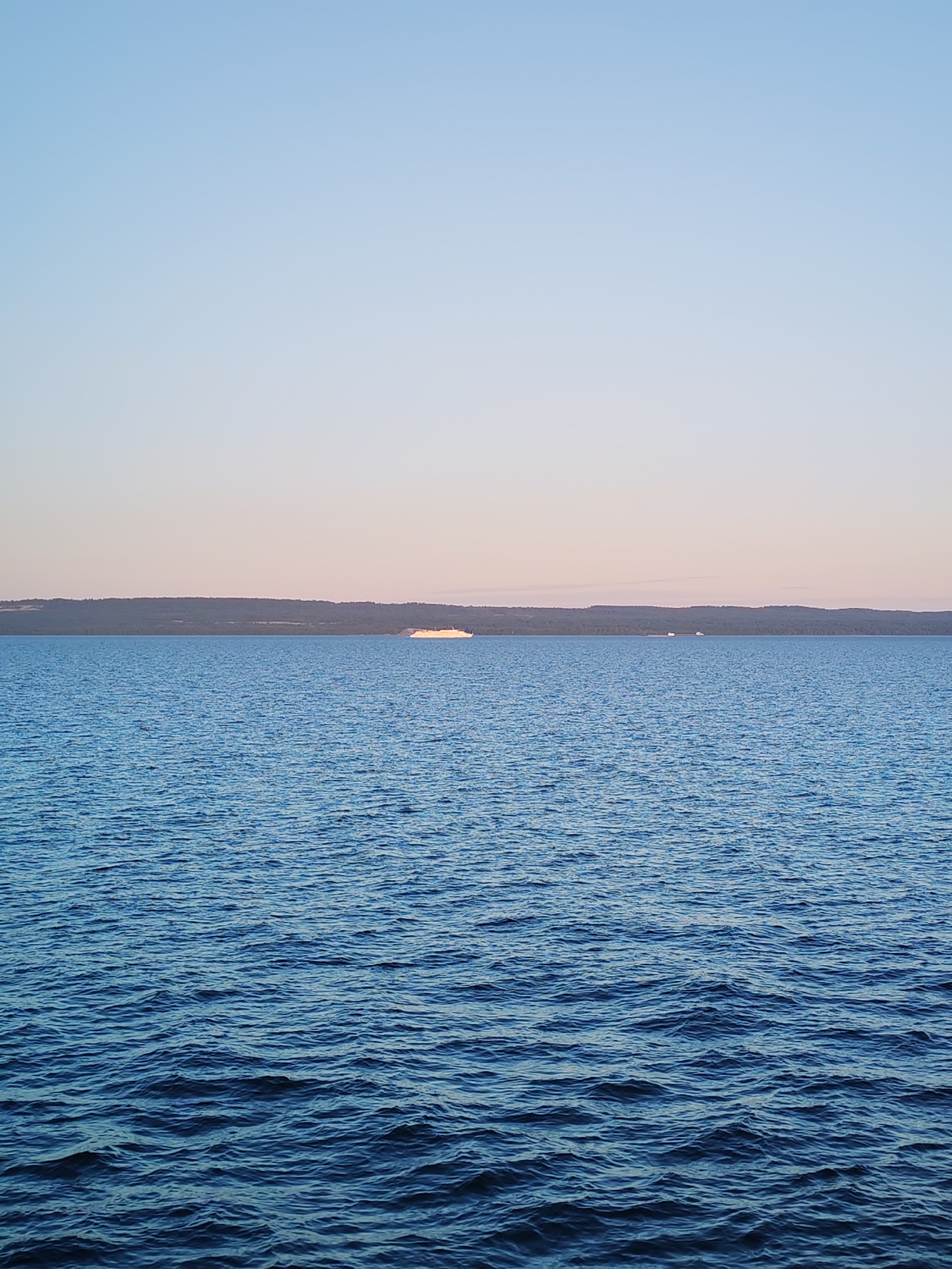 The image captures a serene seascape at dusk or dawn, with the sky transitioning from a pale blue at the top to a soft pinkish hue closer to the horizon. The vast expanse of the water is a deep blue, with gentle ripples reflecting the diffused light. In the distance, a long, tree-covered shoreline stretches across the frame. A white passenger ferry or large boat is visible on the water, approximately in the middle of the frame, with a smaller vessel to its right. The overall scene conveys a sense of tranquility and natural beauty. The image is a photograph taken in Kakkarovo, Russia.