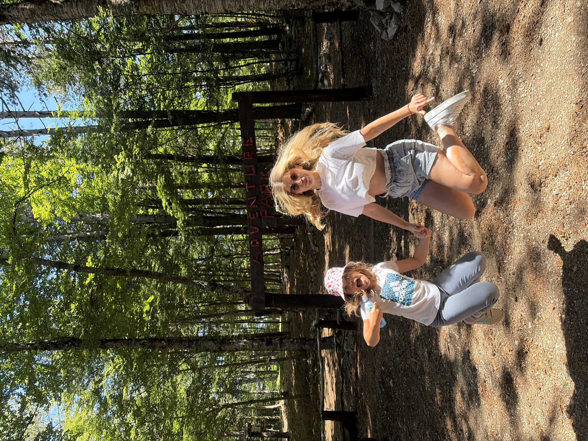 In Mali Bostur, Montenegro, a bright daylight scene captures an adult female and a young girl mid-jump within a wooded adventure park. Both individuals are airborne, holding hands, and looking towards the camera with expressive faces.
The adult female, positioned on the right, has long blonde hair dramatically fanned upwards by the jump. She wears sunglasses, a white cropped t-shirt, light blue denim shorts with a ruched elastic waist, and white sneakers. Her arms are slightly outstretched.
The young girl, on the left, wears a white bucket hat with a red pattern, a white t-shirt featuring a blue and black Adidas logo, light grey sweatpants, and light-colored sneakers with pink accents. Her mouth is open in an exclamation or laugh, and she holds a small clear plastic water bottle in her left hand.
Behind them, a rustic wooden sign stands on two posts, bearing the red text "ADVENTURE PARK". The environment is a forest with numerous tall trees featuring green foliage and light-colored bark. Patches of clear blue sky are visible through the canopy. The ground is a mixture of light brown gravel and dirt, illuminated by dappled sunlight that creates distinct shadows. Additional wooden structures, likely part of the adventure park, are visible in the deeper background to the left.