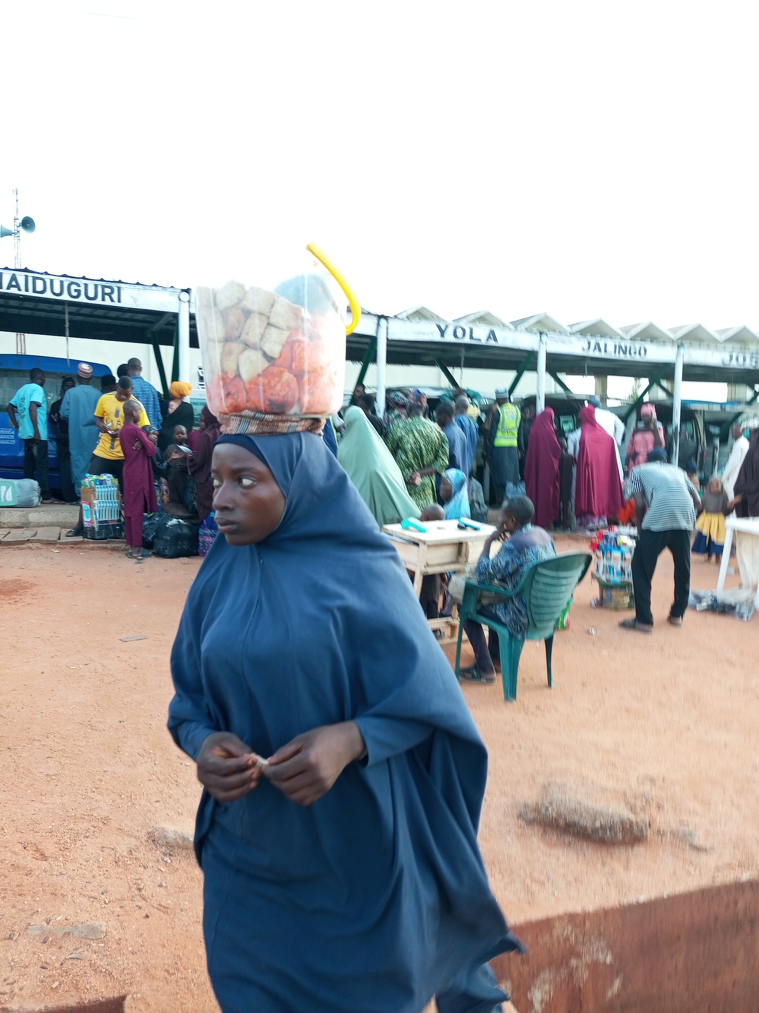 A young woman, seen from the chest up and facing left, walks across a dusty area. She wears a dark blue hijab and a matching, loose-fitting garment that drapes over her shoulders and chest. On her head, she balances a clear plastic bucket filled with what appears to be food items, including some reddish spheres and lighter-colored cubes, topped with a yellow handle. Her hands are near her chest, and she holds a small, light-colored object, possibly a piece of food, between her thumb and forefinger.
In the background, a bus station with signage indicating destinations like "MAIDUGURI," "YOLA," and "JALINGO" is visible. Several people are gathered around buses and under a covered shelter. Some are standing, others are seated. The ground is a reddish-brown, sandy dirt. The sky is overcast and pale. The overall scene suggests a busy transportation hub.