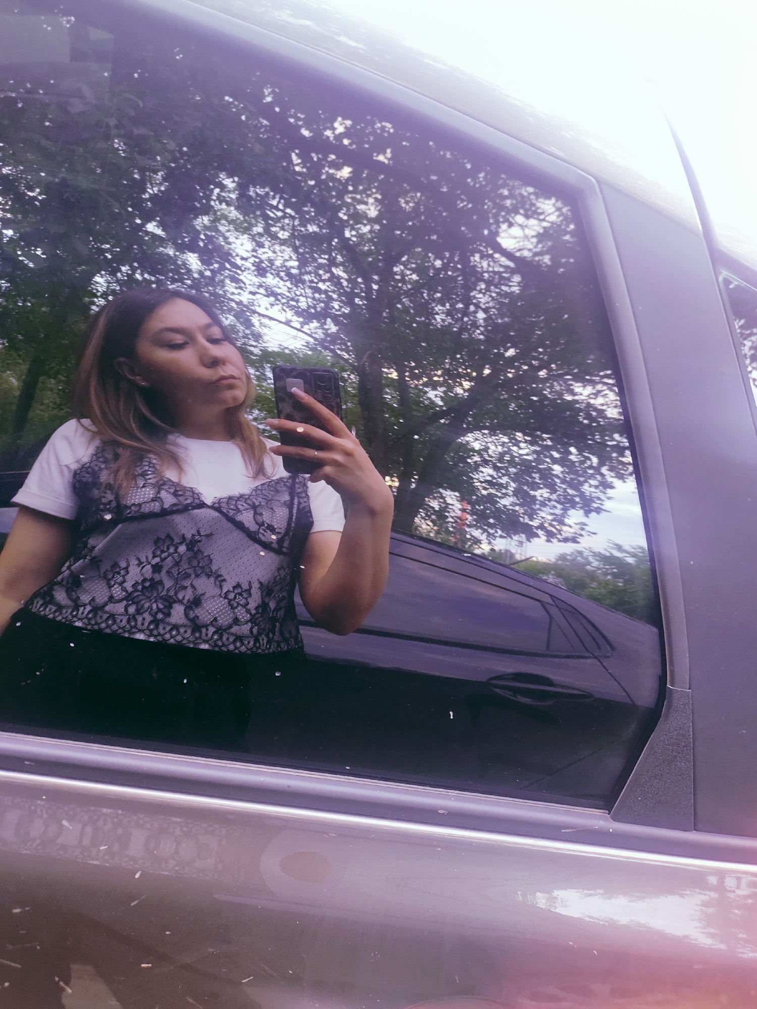 A young woman with brown hair is reflected in the window of a car, taking a selfie. She is wearing a white t-shirt with a black lace top over it and black pants. The reflection shows trees and the sky in the background, suggesting an outdoor setting. The image is taken at eye-level, capturing the woman from the chest up. There are no discernible city landmarks or specific indicators of the location in Chaikovsky, Russia.