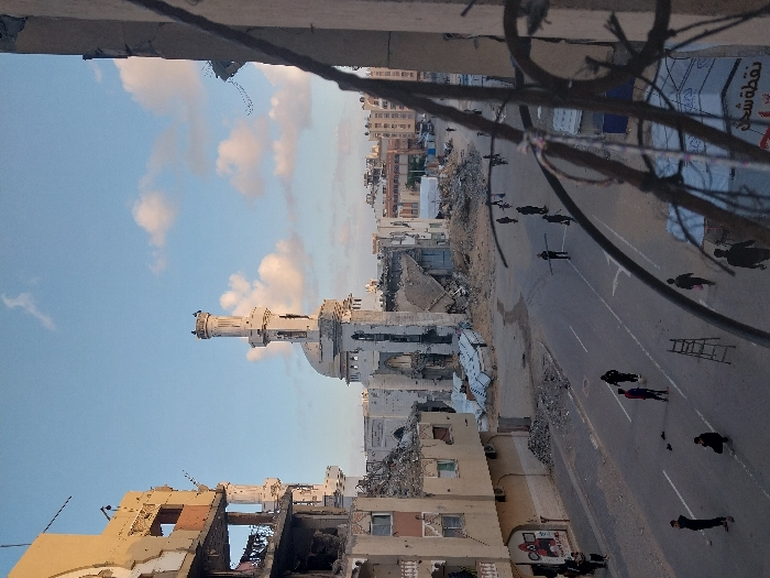 This detailed image depicts a severely damaged urban landscape in Khan Yunis, Palestinian Territory, under a partly cloudy, light blue sky. The central focal point is a significantly damaged mosque with a white dome and a tall, multi-tiered minaret that remains largely intact, though the mosque's main structure is heavily dilapidated with large sections of its walls missing and rubble visible inside.
To the left of the street, a multi-story building, likely a former residential or commercial structure, exhibits extensive damage, with its facade partially collapsed and interior elements exposed. Debris is scattered on the ground below this building. On one section of a wall, a faded "Squid Game" graphic and Arabic text are visible. Other damaged buildings line the street further into the distance.
The asphalt street, marked with white lane lines, is largely clear of vehicles but populated by approximately 15-20 individuals, predominantly males, dressed in dark clothing, walking in various directions. Several people are carrying long, pole-like objects, possibly salvaged materials. A metal A-frame ladder lies horizontally in the middle of a lane. Small piles of rubble, dirt, and debris are scattered across the street and sidewalks.
The foreground on the right side of the image is partially obscured by tangled, rusted rebar and wires, suggesting the photograph was taken from within a damaged structure. Some makeshift tarpaulin shelters are visible near the bottom right. The overall scene conveys widespread destruction and ongoing human movement amidst the rubble.
