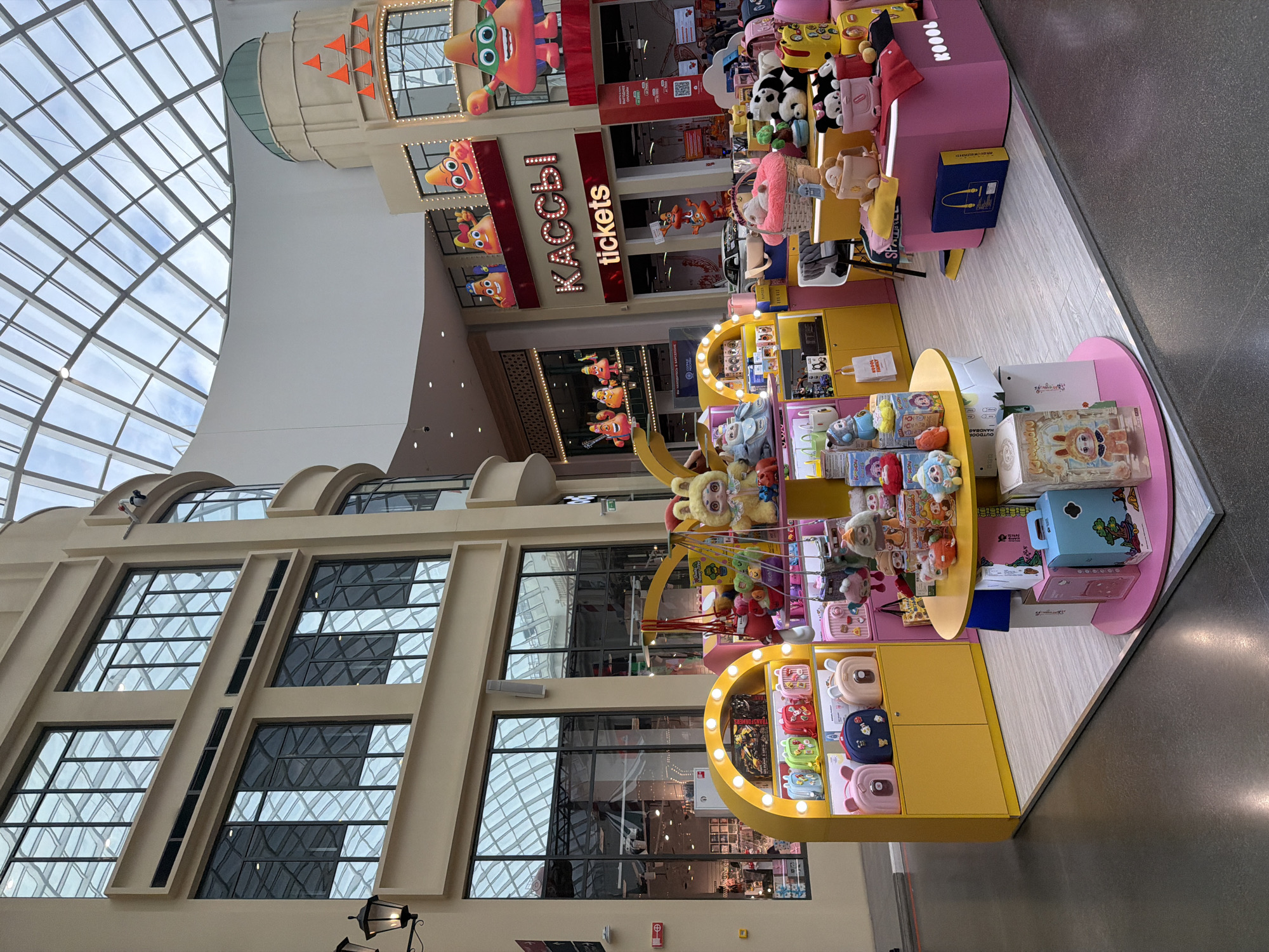 This image shows a store selling plush toys and bags, likely in a shopping mall. The store has bright yellow and pink displays featuring a variety of stuffed animals and small backpacks with cartoonish designs. Some of the plush toys are panda-shaped, and others are more abstract characters. The backpacks come in various colors and have animal-like faces or other playful graphics.
Behind the toy display, a building facade with large windows and a glass ceiling can be seen, suggesting an indoor, well-lit environment. There are signs with text visible, including "KOOC," "tickets," and what appears to be "TRANSFORMERS" on one of the displayed items.
The overall impression is a vibrant and cheerful retail space, likely aimed at children or young adults, as indicated by the colorful and whimsical merchandise. The lighting suggests it is daytime. There are no people clearly visible in the foreground.