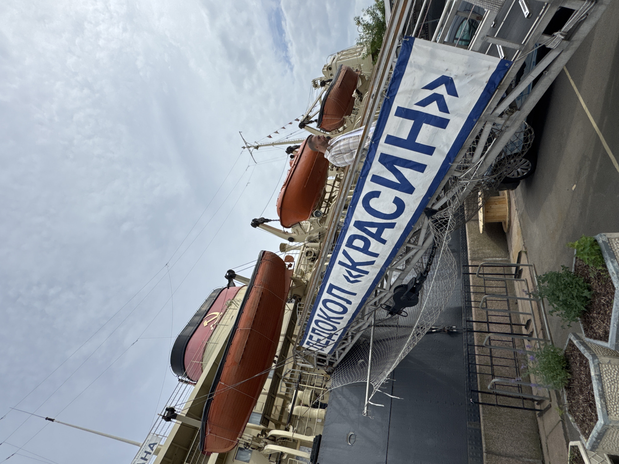An outdoor, low-angle, eye-level shot shows a portion of a large, grey-hulled ship docked in Saint Petersburg, Russia. The ship has a tall, red-and-black funnel adorned with a golden hammer and sickle emblem. Several orange lifeboats are visible, suspended by davits. A metal gangway extends from the ship to the dock, and a large white banner with blue Cyrillic lettering, reading "ЛЕДОКОЛ «КРАСИН»" (Icebreaker "Krasin"), is affixed to it. A man in a plaid shirt and white t-shirt is walking up the gangway. The background features a cloudy, pale blue sky. In the foreground, a metal fence surrounds some planters with greenery. The ship's hull is a dark grey, and the dock area appears to be paved.