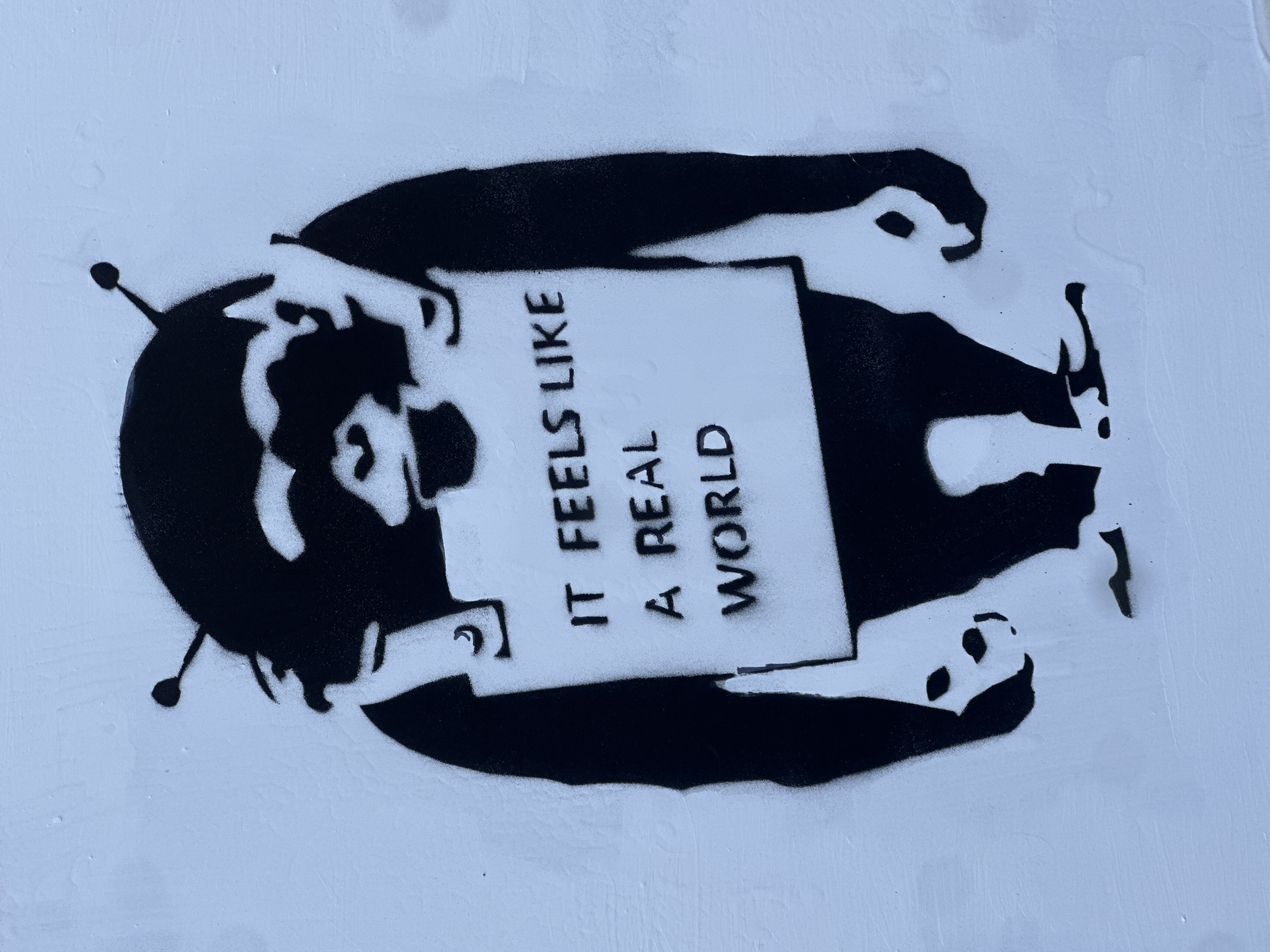 The image shows a black and white stencil of a person holding a sign with the text "IT FEELS LIKE A REAL WORLD." The style resembles the work of street artist Banksy. The location in the city of London, United Kingdom is not discernible from the image alone.