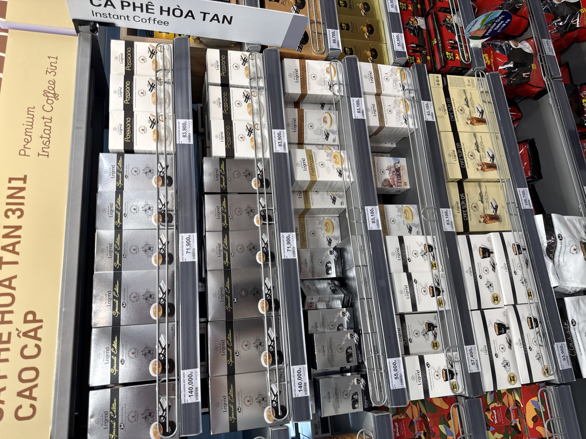 The image shows a retail store display of various coffee products in Nha Trang, Vietnam. The shelves are stocked with boxes of instant coffee from the brand "Legend" and other brands. Prominently displayed are "Legend Special Edition", "Legend Passiona", "Legend Cappuccino", and "Legend Americano" coffee varieties. Prices are listed in Vietnamese Dong (₫). For example, a "Legend Special Edition" box is priced at 140,000₫, and "Legend Passiona" is 83,900₫. The store environment appears to be a supermarket or convenience store with well-lit aisles and organized product placement. The signage at the top indicates a focus on "Premium Instant Coffee 3in1" and "CÀ PHÊ HÒA TAN" (Instant Coffee). The lower shelves display larger bags of coffee and what appear to be gift items. One sign mentions "Popular Gift Items" and shows images of coffee cups. The overall impression is a commercial setting dedicated to the sale of coffee products.