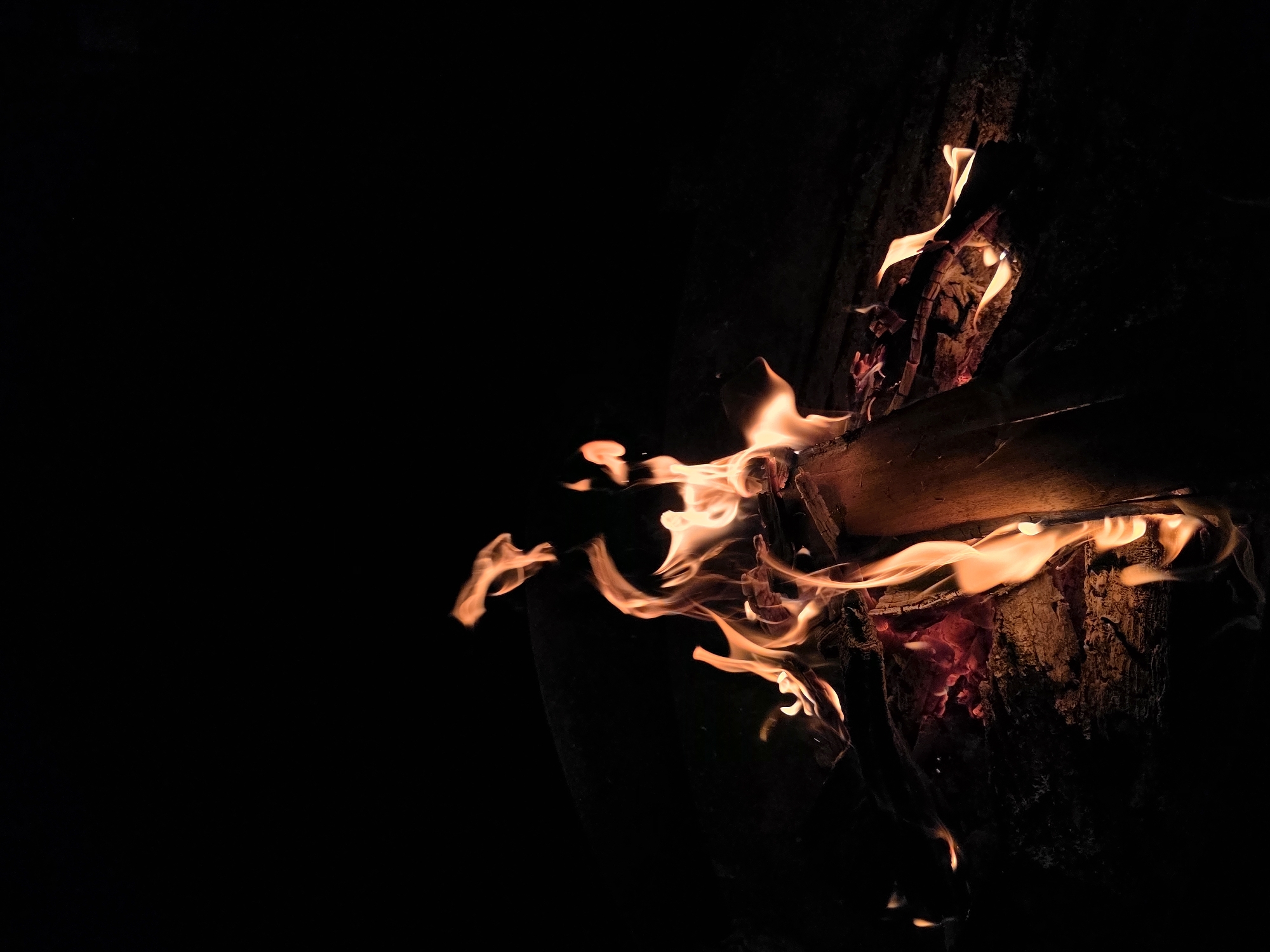 An outdoor campfire burns against a dark background. The flames are a vibrant orange and yellow, casting a warm glow on the burning logs and embers. The wood appears to be a mix of smaller branches and larger logs, some of which are splintered and partially consumed by the fire. The ground beneath the fire is rough and uneven, suggesting a natural setting. The overall impression is one of a cozy and inviting scene, perhaps in a backyard or a campsite. The image does not provide any specific information about the city of Строитель-нефтяник Снт, Russia.