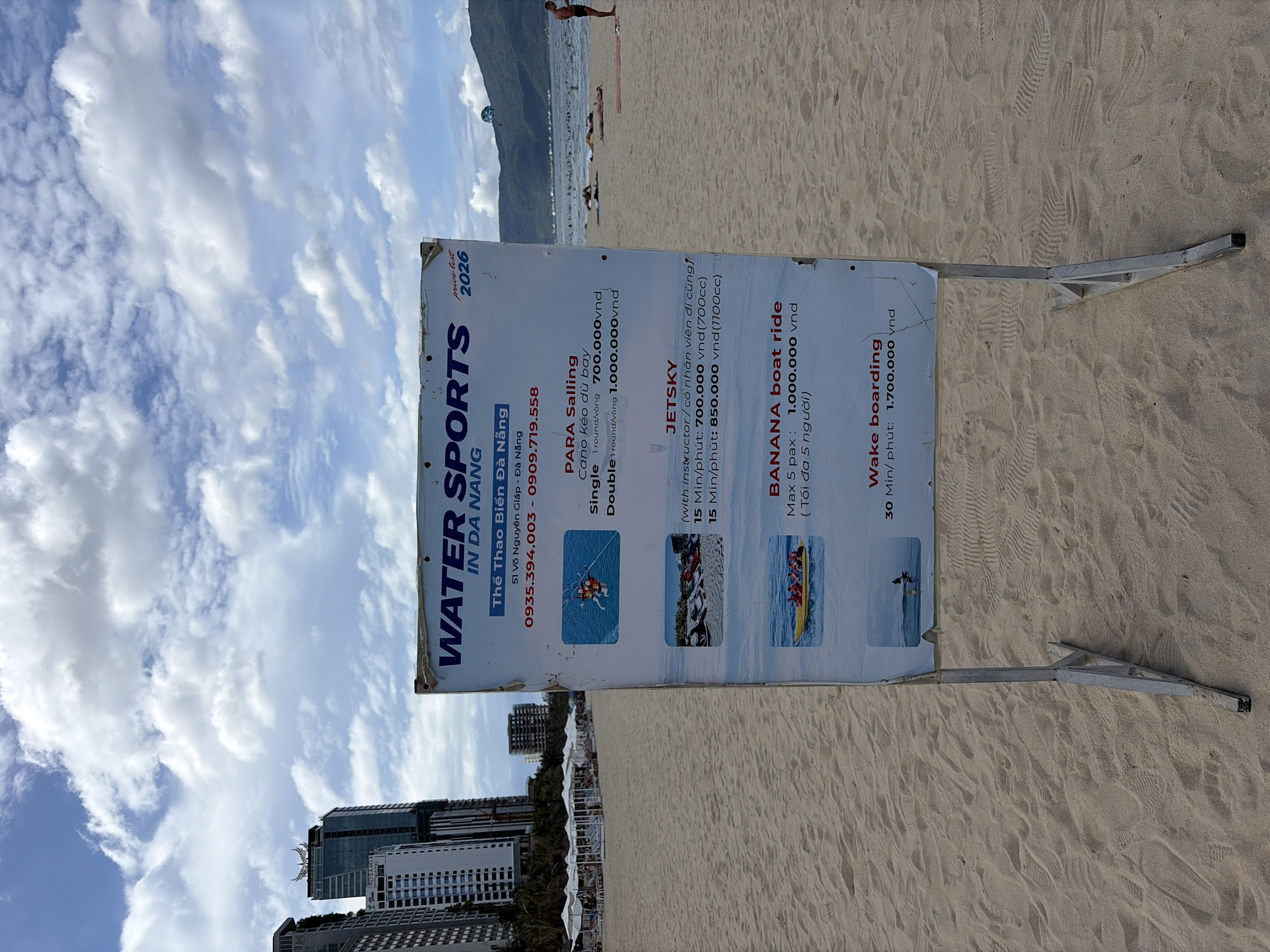 A weathered white sign displaying a price list for water sports stands on a sandy beach in Da Nang, Vietnam. The sign, positioned centrally, is supported by two metal legs. In the foreground, the sandy beach stretches out, marked by footprints. In the background, the ocean is visible with a few people and a distant mountain range under a partly cloudy sky. The sign features text and images detailing services such as "PARA Sailing," "JETSKY," "BANANA boat ride," and "Wake boarding." The list includes prices in Vietnamese Dong (vnd) and durations, with some text in Vietnamese. The overall scene depicts a typical day at a beach resort offering recreational activities.