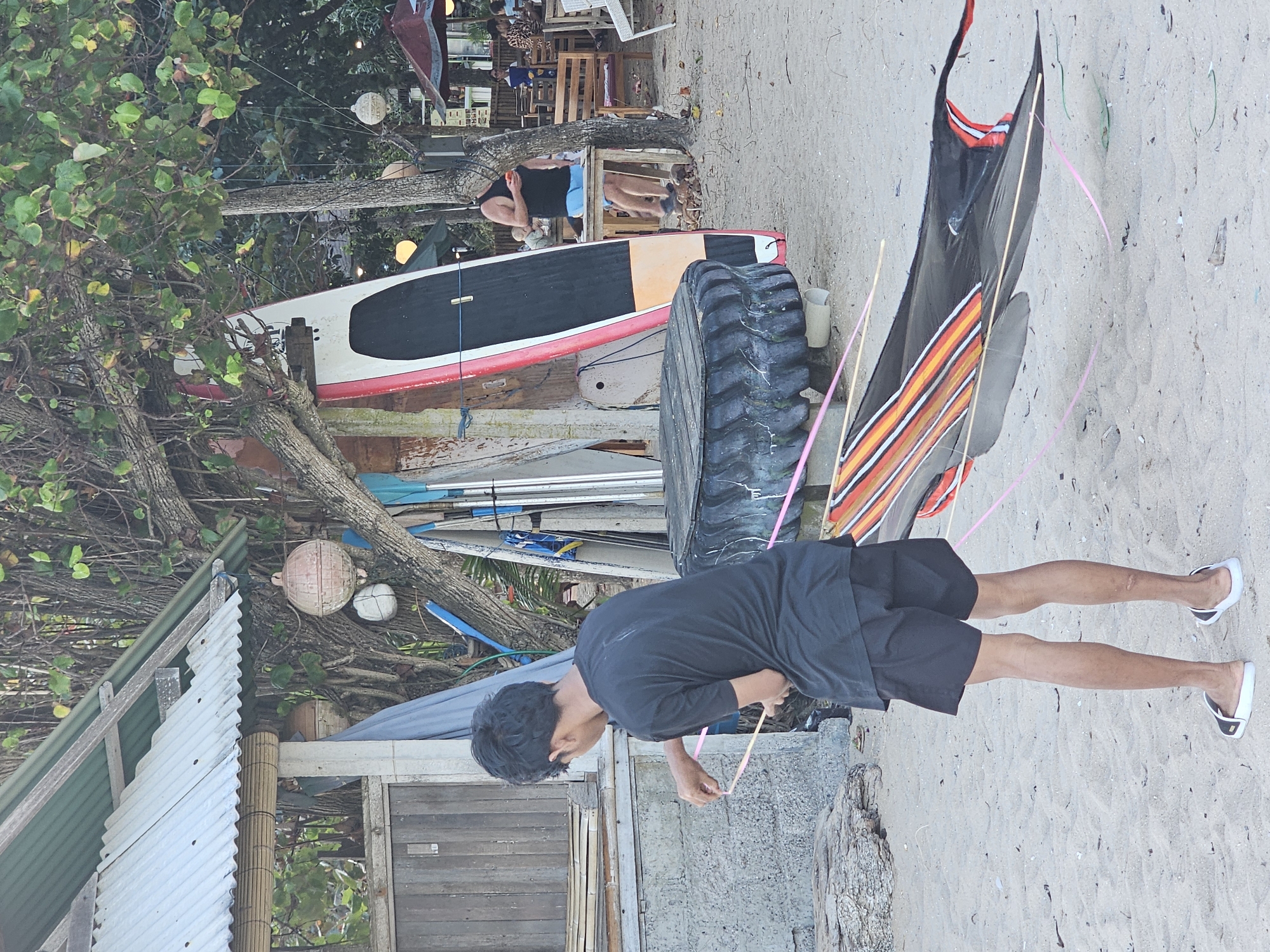 The image depicts a beach scene in Denpasar, Indonesia. In the foreground, a young male wearing a black t-shirt and black shorts is standing on the sand, holding lines attached to a large, black kite with orange and red accents that lies on the ground. To his right, there is a large tire serving as a makeshift table. In the background, several surfboards are propped up against a building structure. A man in a black tank top and blue shorts is standing near a table and chairs in a seating area. Further in the background, more seating areas and umbrellas are visible, suggesting a cafe or restaurant. Trees with foliage are present throughout the scene, indicating a tropical environment. The overall setting is a beach with elements of recreational activities and relaxation.