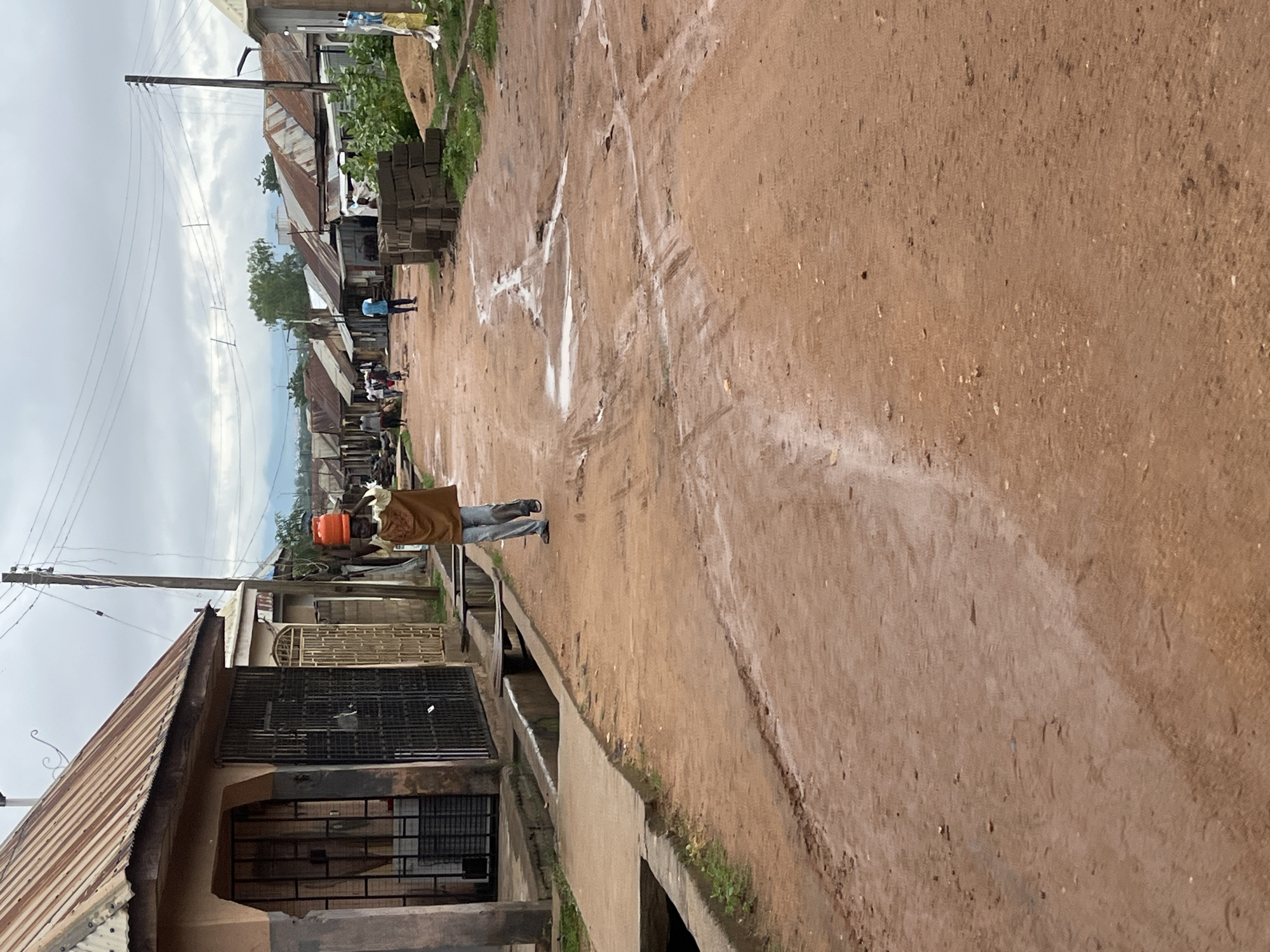 A person walks down a dirt road in Jalingo, Nigeria, carrying two orange containers on their head. The person is wearing a brown vest over a light-colored shirt and light-colored pants. The road is muddy with puddles of water. On the left side of the road, there are buildings with corrugated metal roofs and barred windows. On the right side of the road, there are more buildings, some made of corrugated metal, and a stack of bricks. In the distance, there are more buildings and some vegetation. The sky is overcast.