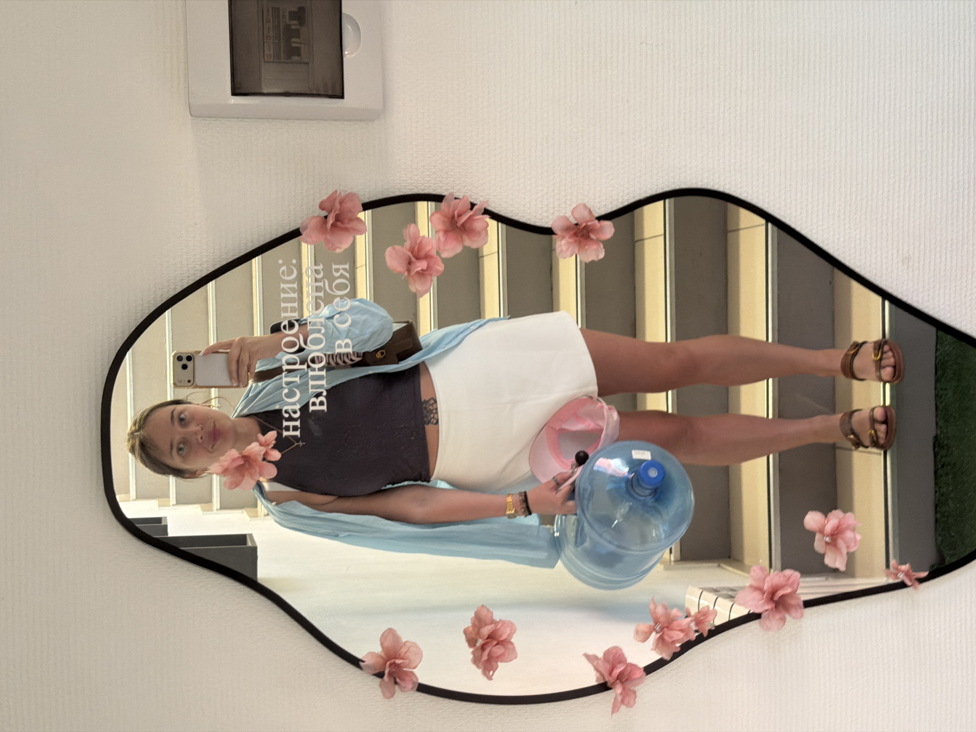 An indoor mirror selfie captures a young woman posing in what appears to be a building interior in Chaikovsky, Russia. The woman, likely in her 20s or 30s, has light brown hair pulled back. She is dressed in a dark crop top, a white mini skirt, a light blue unbuttoned long-sleeve shirt, and brown flat sandals. She holds a light-colored smartphone in her right hand to take the photo and a large, clear blue water jug in her left hand. A pink baseball cap is visible near her left hip, and a small tattoo is on her left lower back.
The mirror has an irregular, wave-like black frame and is decorated with multiple pink artificial flowers attached to its edges. The reflection in the mirror reveals a staircase with light-colored risers and darker treads, along with rectangular architectural elements that resemble windows. A small patch of green, possibly grass, is visible in the lower right corner of the reflection.
The wall surrounding the mirror is white and textured, with an electronic device, possibly an intercom or control panel, mounted above it. Visible text on the mirror, written in Cyrillic and appearing backward due to the reflection, reads: "настроение: Влюблена в себя." This translates to "Mood: In love with myself." The scene is brightly lit, suggesting it is daytime. The woman gazes towards the camera with a neutral expression.