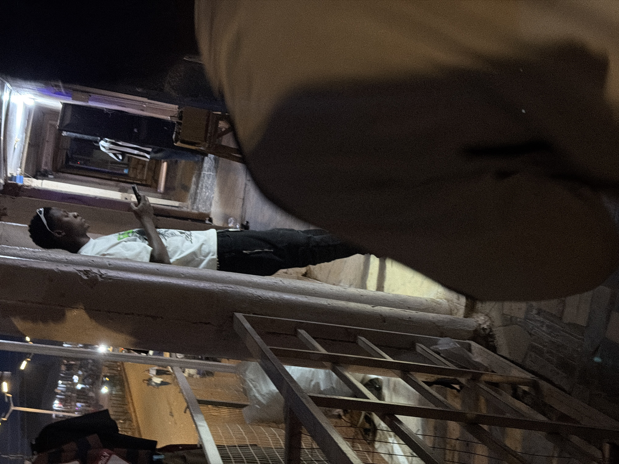 In Bukuru, Nigeria, at night, a young Black man wearing a white t-shirt and dark pants stands on a balcony, looking at his phone. He has sunglasses pushed up on his forehead. Behind him, the walkway of the building continues, with another person in a black and white striped shirt visible further down. A wooden railing separates the balcony from the street below, where cars and pedestrians are present. Clothing is hanging on a rack to the left of the frame. The lighting is dim, suggesting an urban environment at night.