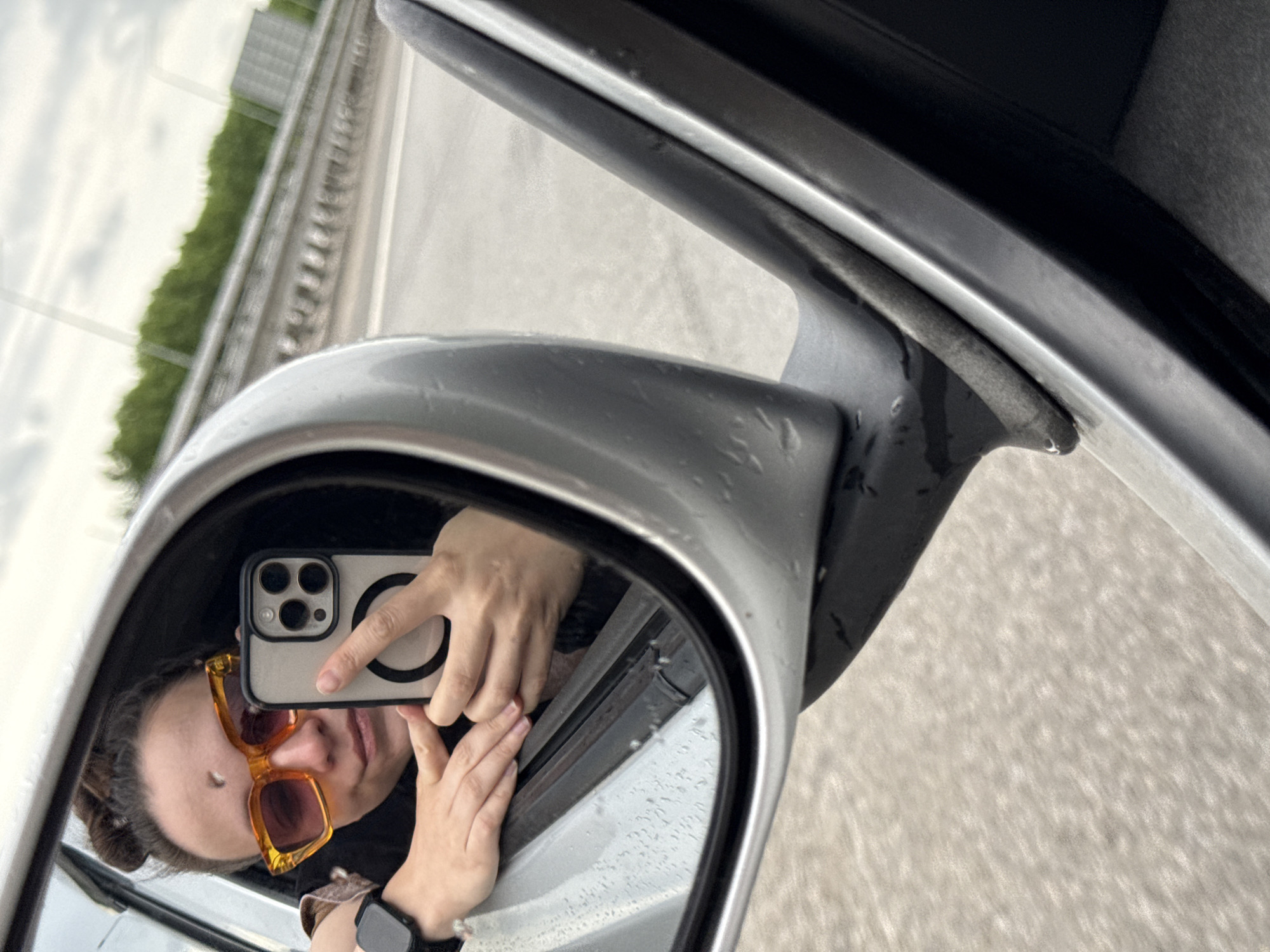 A person is taking a selfie in a car's side mirror. The person has sunglasses and a watch. The environment appears to be a road with a highway divider in the background. Based on the prompt, the location is assumed to be in the city of Shali, Russia.