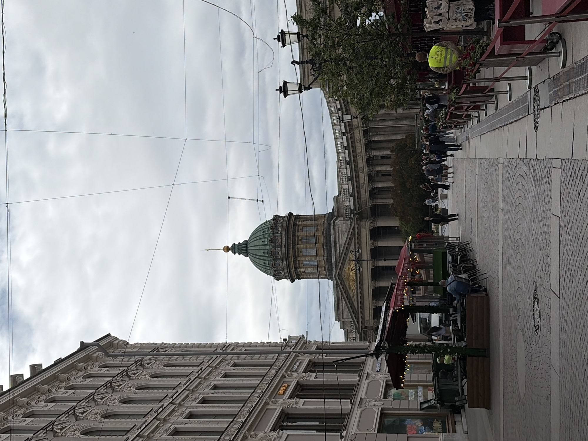 The image depicts a street scene in Saint Petersburg, Russia, on an overcast day. In the foreground, an ornate building with numerous windows and decorative elements lines the street. Beyond this, a grand domed structure, likely a cathedral or significant architectural landmark, is visible. Several people are present, some walking along the paved sidewalk and others gathered near outdoor seating areas or kiosks. One person in a bright yellow vest stands out, possibly working at an event or providing assistance. String lights are visible on some of the structures, suggesting a festive or commercial atmosphere. The sky is cloudy, indicating a typical Saint Petersburg weather day. The overall impression is one of urban life with a blend of historical architecture and contemporary activity. Visible text includes the word "БАЙКИНА" on the building in the foreground.