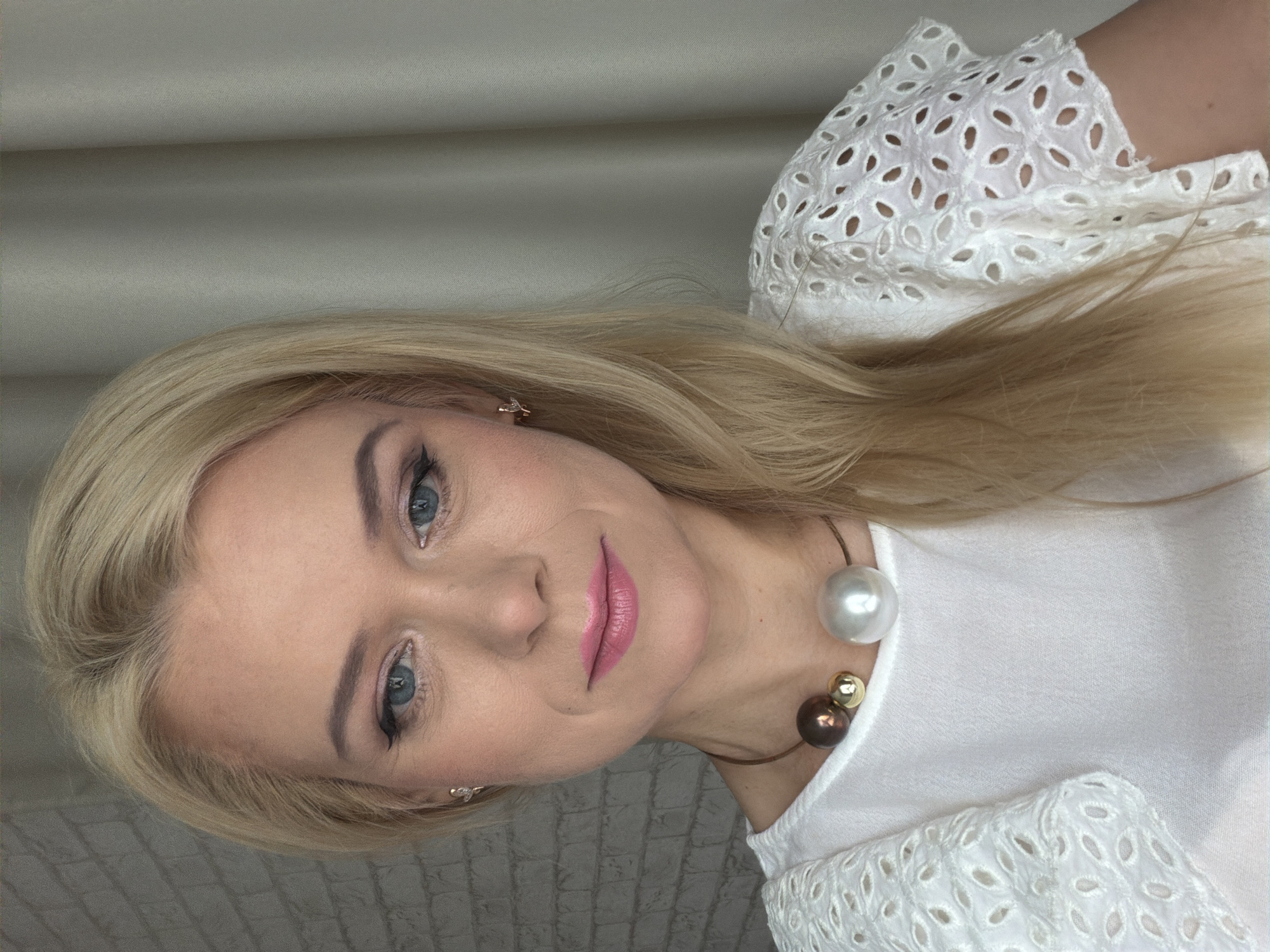The image is a portrait of a blonde woman with blue eyes, wearing makeup and a white, eyelet-detailed shirt. She is also wearing a necklace with large, round beads in white, brown, and gold, and small stud earrings. The woman is looking towards the camera with a slight smile. The background consists of a light-colored, textured wall on the right and a plain, light-colored fabric on the left, which appears to be a curtain or backdrop. The lighting suggests an indoor setting, likely during the daytime. The image is oriented sideways. No text is visible in the image.