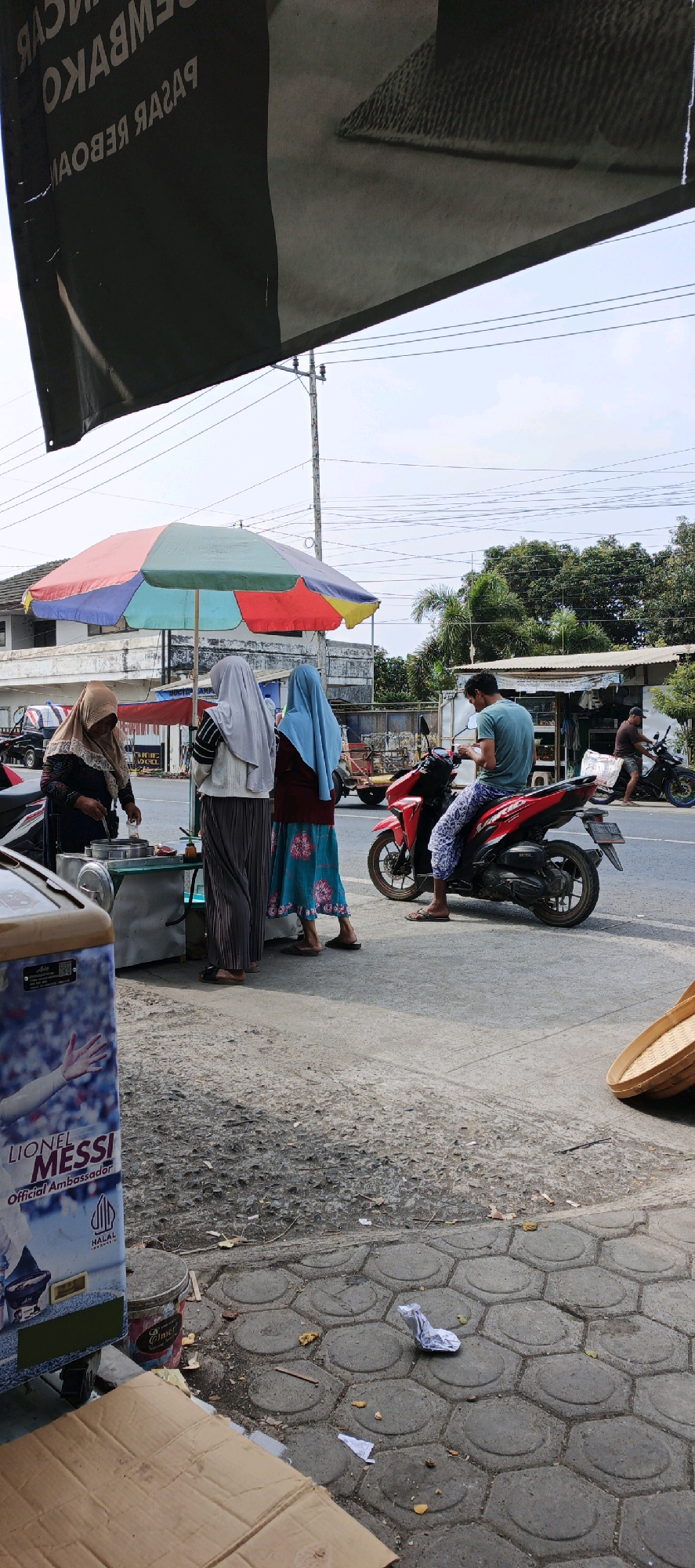 The image depicts a street scene in Kasiyan, Indonesia. A vendor, wearing a brown hijab, is serving food from a cart under a colorful umbrella. Two women, one in a light grey hijab and the other in a blue hijab, are standing near the cart, appearing to interact with the vendor. A man wearing a blue t-shirt and patterned pants is sitting on a red motorcycle, looking at his phone. Another man is riding a black motorcycle in the background. The street is paved with interlocking hexagonal concrete blocks in the foreground, transitioning to asphalt further down. A large banner with text, possibly advertising, is visible at the top of the image. In the foreground, a blue cooler or refrigerator with an image of Lionel Messi is present, along with some discarded cardboard and a paint can. The environment is outdoors with trees and buildings visible in the background, under a cloudy sky.