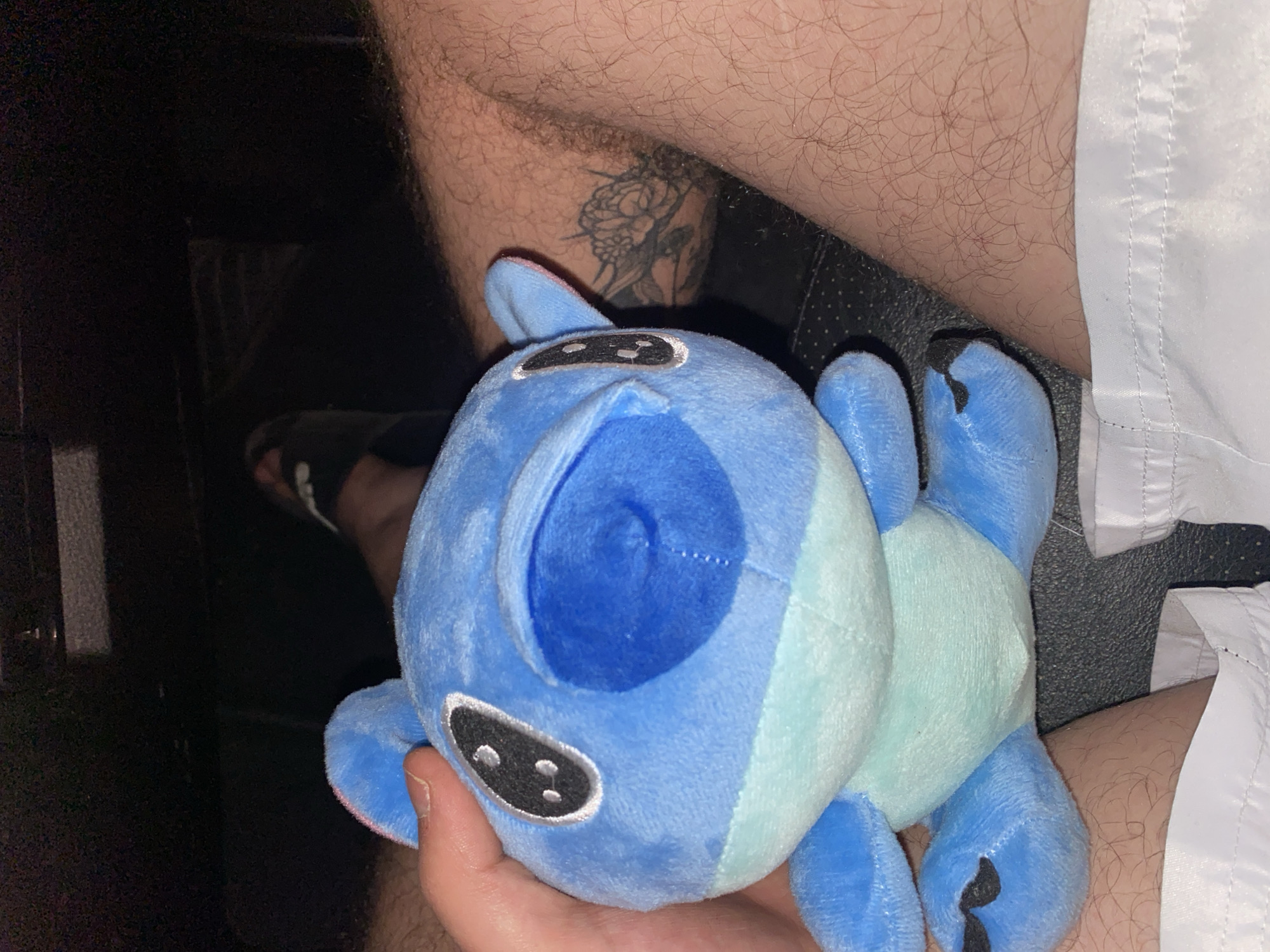 The image shows a close-up of a person's legs and a plush toy, likely in a dimly lit indoor setting. A hand is holding a blue plush toy, which appears to be a character with large black eyes and a light blue stomach. The person's legs are visible, with one leg adorned with a tattoo of a dragon-like creature. The person is wearing white shorts. The background is dark, with hints of what might be car interior elements. The context of Yasen, Bulgaria, is not discernible from the visual information in the image.
