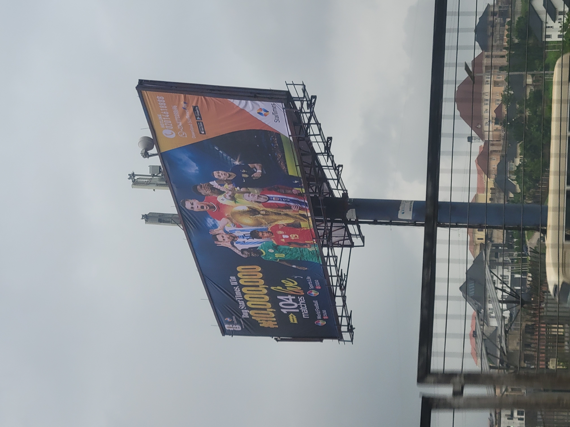 The image shows a billboard advertising StarTimes, a pay-TV operator, with the slogan "Buy StarTimes, Win #10,000,000, 104 live matches." The billboard features images of several famous football players, including Lionel Messi, Cristiano Ronaldo, Sadio Mané, and Kylian Mbappé. The billboard also displays a hotline number and an invitation to download the StarTimes app from the App Store or Google Play.
The billboard is mounted on a metal structure, which is supported by a tall pole. In the background, there are several residential buildings with various roof styles, suggesting an urban or suburban setting. The sky is overcast, indicating a cloudy or possibly rainy day. The lower part of the image shows a metal fence or barrier, through which the buildings in the background are partially visible.
This billboard is likely located in the city of Aso, Nigeria, given the context of StarTimes' operations and the visual cues that are consistent with Nigerian urban landscapes.