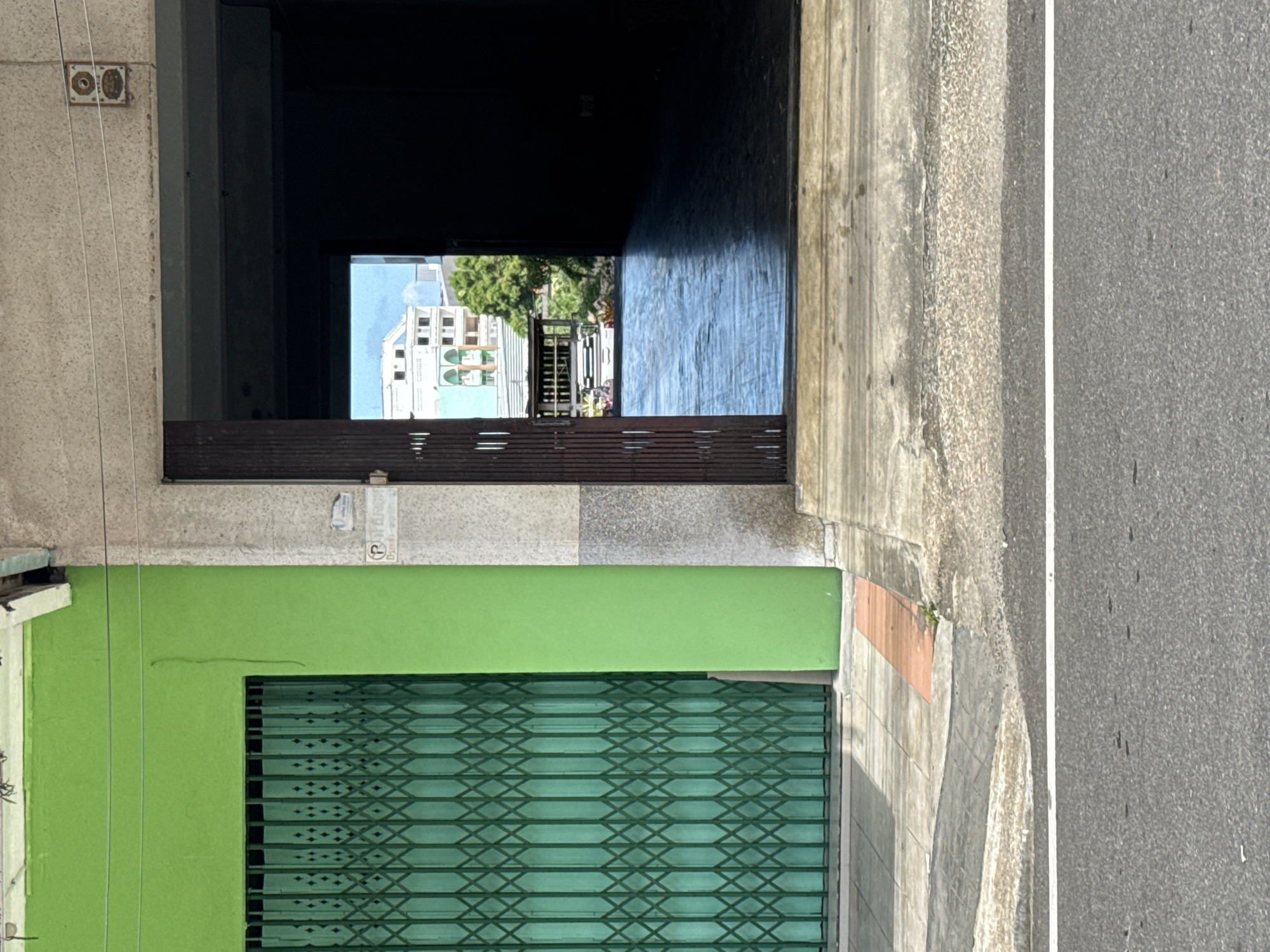 The image depicts a street scene in Ranong, Thailand, focusing on the entrance to a building and its exterior. On the left is a bright green building with a closed, dark green, metal accordion gate covering its storefront. The gate has a diamond pattern. Adjacent to the green building, to its right, is a concrete structure with a rough, mottled grey texture. This structure has an open doorway revealing a dark interior space, partially illuminated by natural light from beyond. Through the open doorway, a sunlit scene of a courtyard or pathway is visible, with a reflective, wet-looking surface. In the distance, a light-colored building with green accents and a white rooftop structure can be seen, along with trees. The ground in front of the buildings consists of concrete sidewalks and a section of asphalt road with a white stripe. The overall impression is one of an urban environment, possibly a commercial area, captured on a bright day.