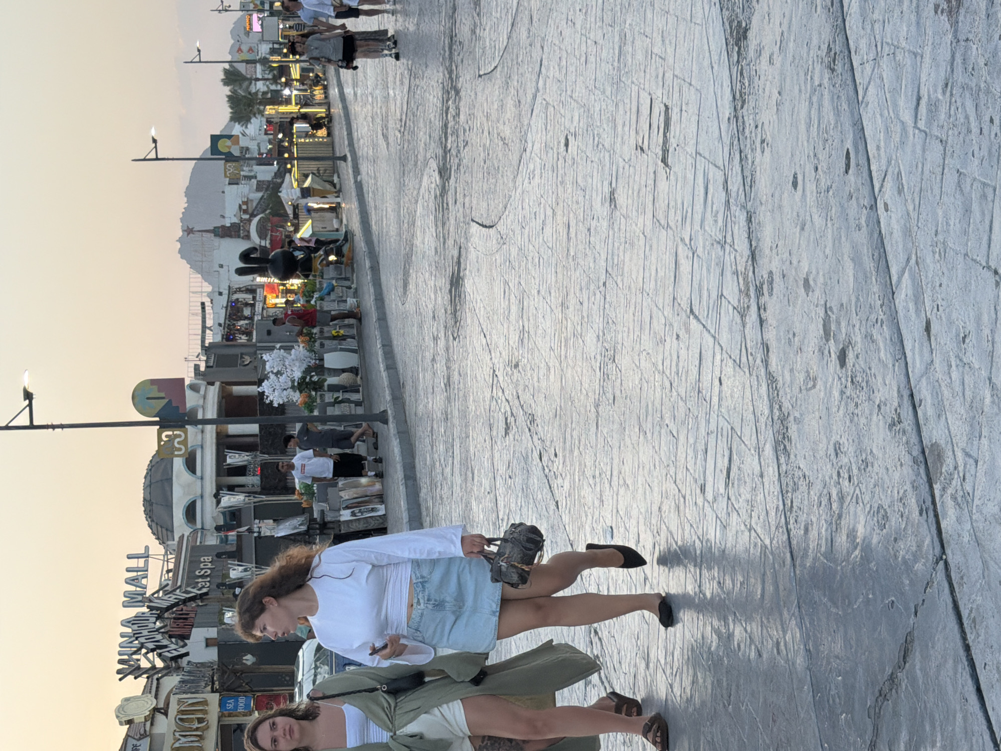 This image depicts a street scene in Sharm El Sheikh, Egypt, likely during sunset, given the warm, soft lighting. Two young women are walking on a patterned cobblestone street. The woman on the left has dark hair and is wearing a white tank top and shorts, with a light green unbuttoned overshirt. The woman on the right has curly brown hair and is wearing a white long-sleeved crop top and a light blue denim skirt. She is carrying a small, dark-colored handbag.
In the background, several shops and buildings line the street, adorned with signs including "Sharm Mall" and "El Zohour Mall." There are various decorative elements such as lamps, plants, and what appears to be a large black bunny sculpture. Other people are visible in the background, walking or standing near the shops. The overall atmosphere suggests a commercial and recreational area. The cobblestone pavement is textured and somewhat wet in places, reflecting the ambient light. A lamppost stands prominently on the right side of the street. The sky is a pale orange-pink, characteristic of a sunset.