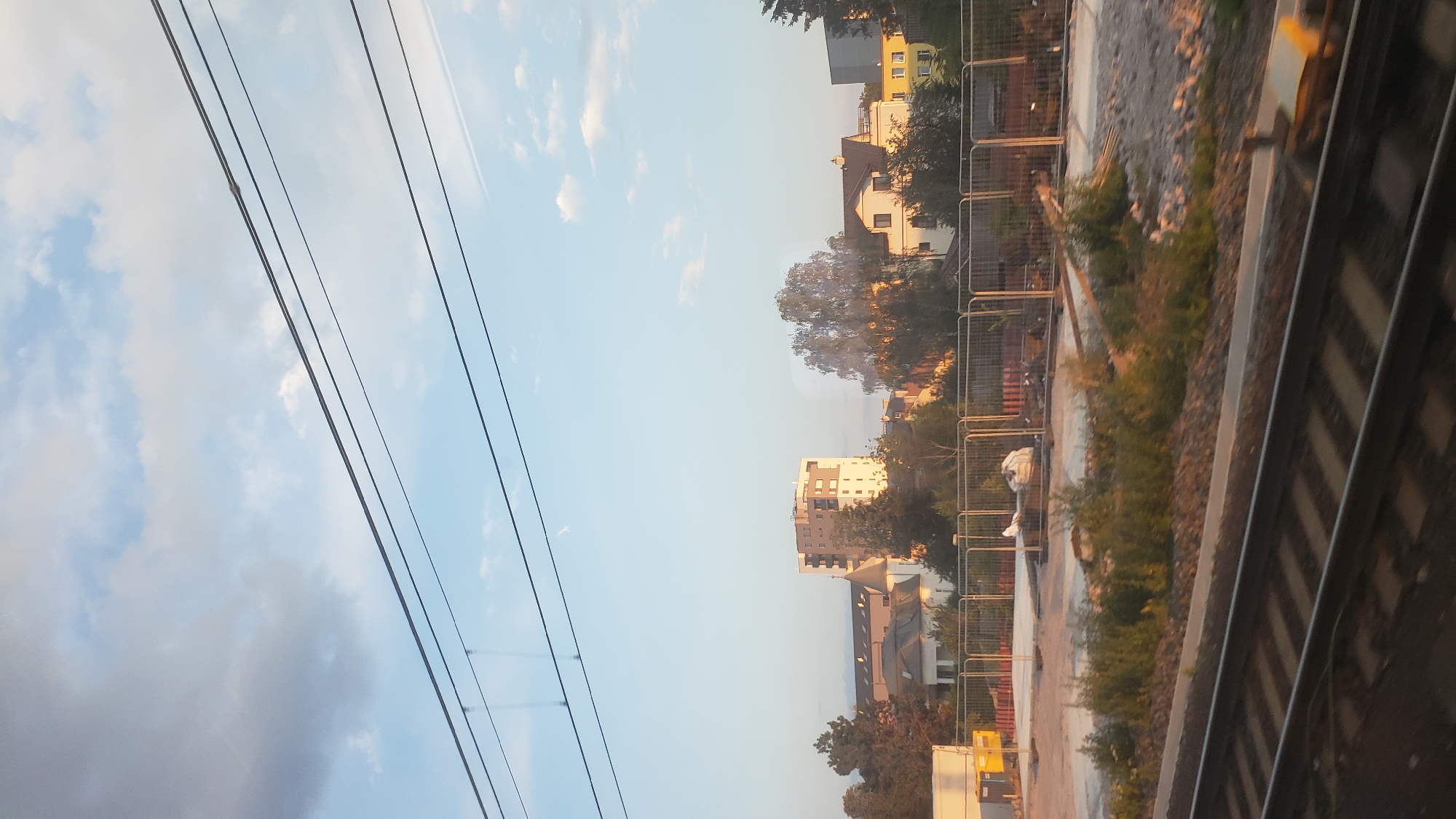 The image depicts a view from a train, likely traveling through Poprad, Slovakia, given the context of the prompt. The foreground features train tracks running horizontally across the bottom of the frame, with ballast and vegetation visible alongside them. In the midground, a fenced-off construction site is present, with some debris and materials visible behind the fence. Beyond the construction site, a cluster of buildings can be seen, including a modern, multi-story building with a light-colored facade, and several other residential structures with pitched roofs. Trees are interspersed among the buildings, with a large deciduous tree prominently featured in the center. The background consists of a clear blue sky with scattered clouds, indicating daytime. The overall scene suggests an urban or suburban landscape viewed during transit. There are no people or animals visible in the image.
