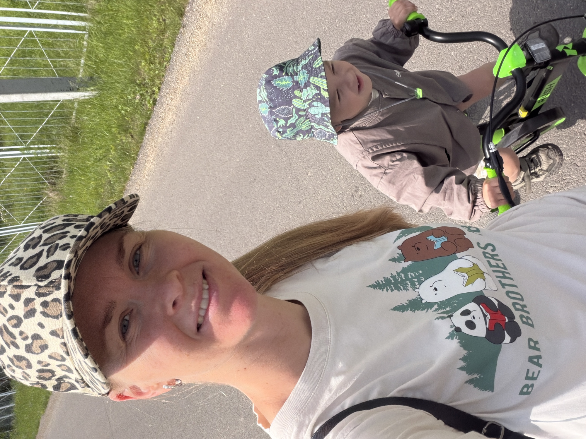 In the city of Kul'dur, Russia, a woman wearing a leopard-print baseball cap and a white t-shirt with a "Bear Brothers" graphic is taking a selfie. She is standing to the left, with her face and upper body visible. To her right, a young child, wearing a green-patterned bucket hat and a brown jacket, is seated on a black and green bicycle. The child is looking down and to the right. Both subjects are on an asphalt road. In the background, there is green grass and a white fence. The lighting suggests it is daytime.
