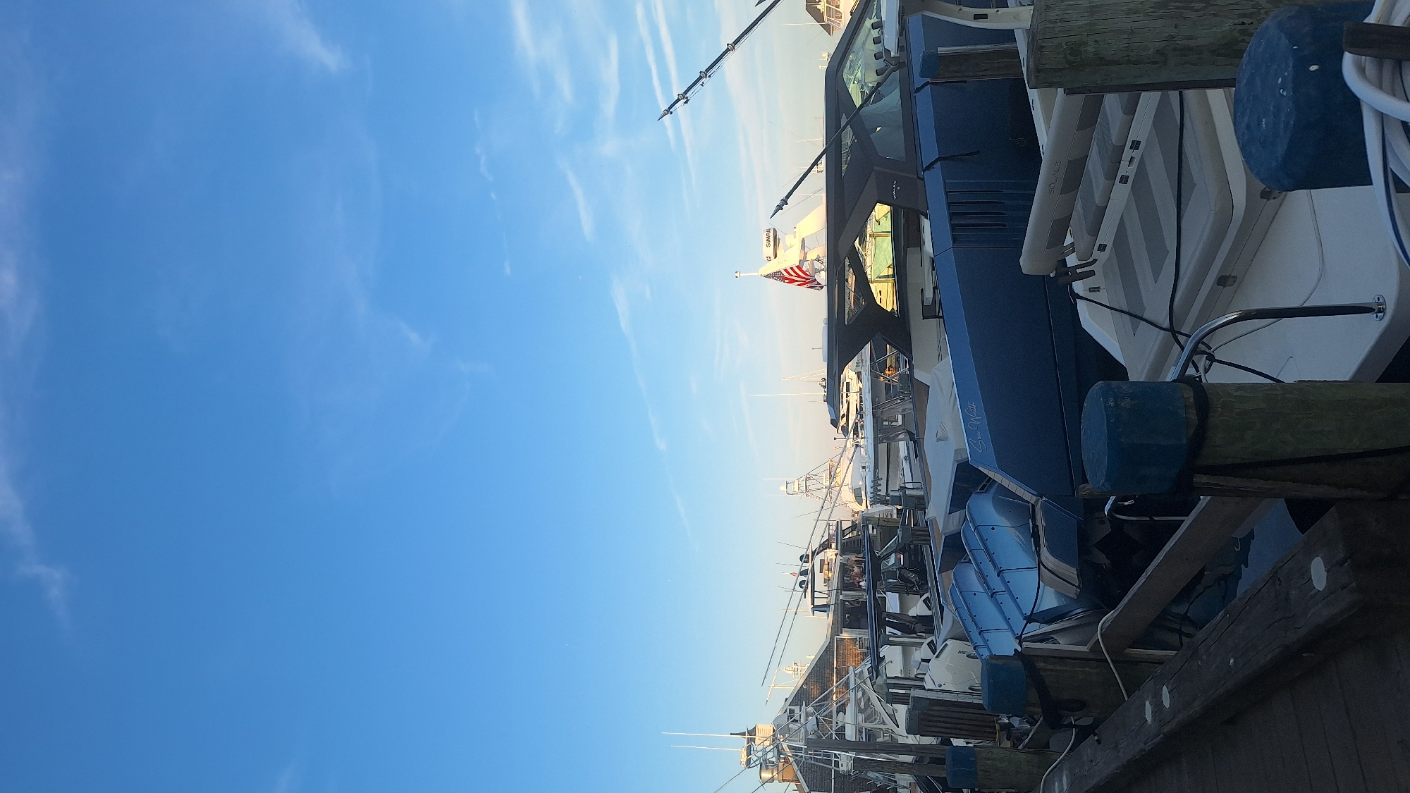 A vertical, medium shot captures a serene scene of boats docked at a harbor in Nantucket, United States of America. The sky, a clear, pale blue with wispy white clouds, dominates the upper portion of the frame. The boats, various sizes and designs, are moored along a wooden dock, their reflections shimmering in the calm water below. The foreground features weathered wooden pilings with blue caps, tied with thick ropes. Several boats are visible, including a sleek, dark blue vessel with a distinctive hull and a white boat with striped seating in the mid-ground. Fishing rods extend upwards from some of the boats. An American flag waves from a mast on a larger boat in the background, adding a touch of national pride to the maritime setting. The overall impression is one of tranquility and a picturesque harbor view.