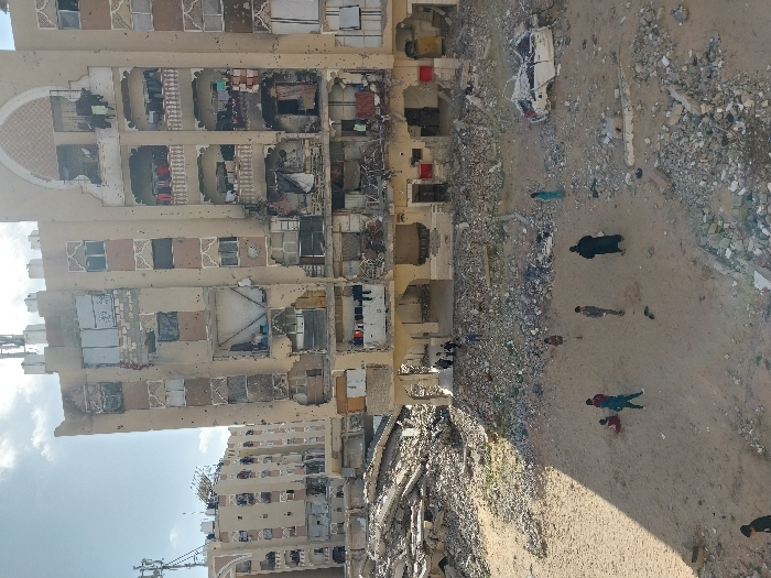 This image captures a severely damaged urban environment in Khan Yunis, Palestinian Territory, under daylight with a partly cloudy sky. The dominant feature is a multi-story, light-colored residential building, occupying the right half of the frame, exhibiting extensive structural compromise. Its facade is perforated with numerous impact marks, and large sections of exterior walls are missing, revealing the interiors of multiple apartments. Clothes are visible hanging from several balconies across different floors. To the left, behind and partially obscured by the foreground building's rubble, another multi-story structure shows similar severe damage and partial collapse.
The ground area in the foreground and midground consists of a sandy, dusty expanse heavily littered with large piles of concrete rubble, rebar, and assorted construction debris. Approximately ten to fifteen individuals, predominantly children, are dispersed across this open, rubble-strewn ground. Actions include standing, with several children and one adult in a dark robe visible, and sitting, with two children observed on the sand in the lower-left. A group of several individuals, including children and adults, is gathered beneath a partially intact archway on the ground floor of the central damaged building. On the right side of the open ground, the heavily mangled, white frame of a vehicle, possibly a car, rests amidst the debris. Communication towers are discernible on the rooftops of background buildings.