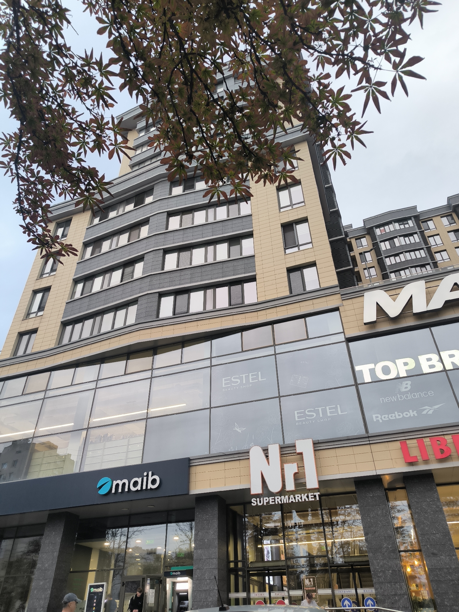 The image shows a modern, multi-story building with a light beige and gray facade, featuring many windows. The lower floors have large glass windows displaying various shop signs. Prominently visible are signs for "maib", "N°1 SUPERMARKET", "TOP BR", "ESTEL BEAUTY SHOP", and "LIB". The "maib" sign is dark gray with a teal circular logo and white text. The "N°1 SUPERMARKET" sign is white and orange. The "ESTEL BEAUTY SHOP" is displayed on the glass facade with a minimalist grey graphic. The "TOP BR" sign is white text on a dark background, with smaller logos for "new balance" and "Reebok" below. Red lettering for "LIB" is also visible. The upper floors of the building have a repeating pattern of windows and beige panels. Trees with reddish-brown leaves are in the foreground, partially obscuring the building. The sky is overcast. The lower entrance area shows glass doors with signage indicating "INTRARE" (Entrance) and "IEȘIRE" (Exit) in Cyrillic and Latin scripts. A person wearing a black hoodie is seen inside the building near the entrance, and another person in a grey cap is visible outside to the left. The scene appears to be a commercial and residential building in Chișinău, Moldova, likely on a street with other businesses.