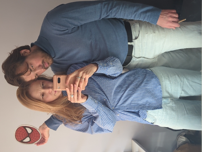 An indoor mirror selfie captures a man and a woman. The woman, with light brown hair, wears a blue and white pinstriped button-down shirt and light blue jeans, holding a rose gold smartphone directly in front of her to take the picture. Multiple rings are visible on her left index, middle, and ring fingers, and on her right ring finger. The man, with dark hair and a beard, stands behind and slightly to her right, wearing a blue turtleneck sweater, light gray jeans, and a black belt. His left arm is around the woman's shoulder, with his right hand extended to hold a red and white Spiderman mask cutout above her head. He also holds a light brown cylindrical object, resembling a cigarette or cigarillo, in his right hand. A simple band-style ring is visible on his left ring finger. The background consists of a plain, light-colored wall, with a white window ledge partially visible on the left side of the frame.