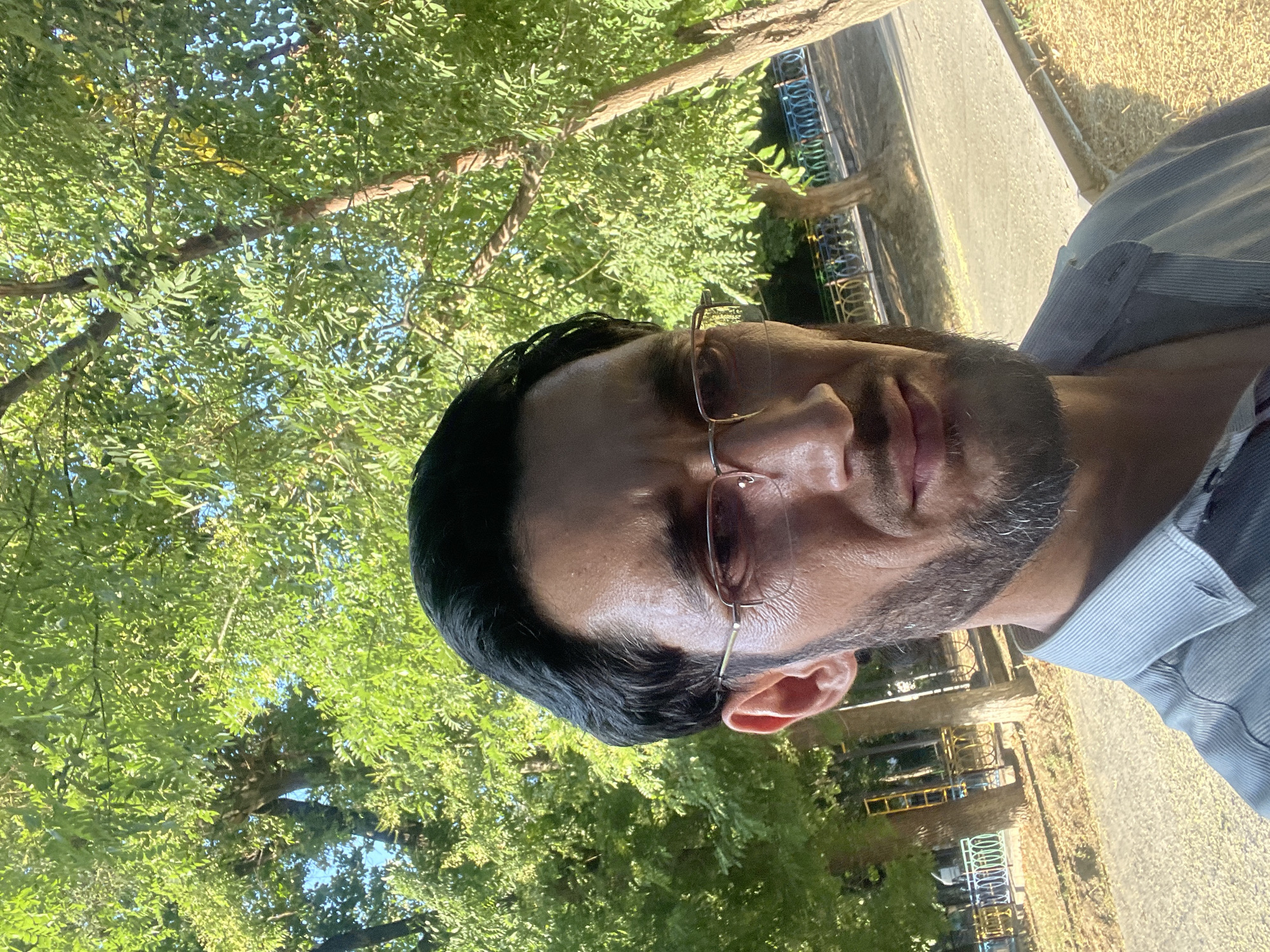 The media file is a selfie of a person with dark hair and stubble, wearing glasses and a light blue striped shirt. The person is looking at the camera with a neutral expression. The scene is outdoors, with lush green trees and foliage in the background, suggesting a park or garden setting. The lighting indicates it is daytime, likely morning or afternoon, with sunlight filtering through the leaves. A paved walkway is visible in the background, along with some industrial-looking structures. There is no visible text in the image. The overall impression is a casual outdoor moment captured in a natural environment. The location context is Tashkent, Uzbekistan.