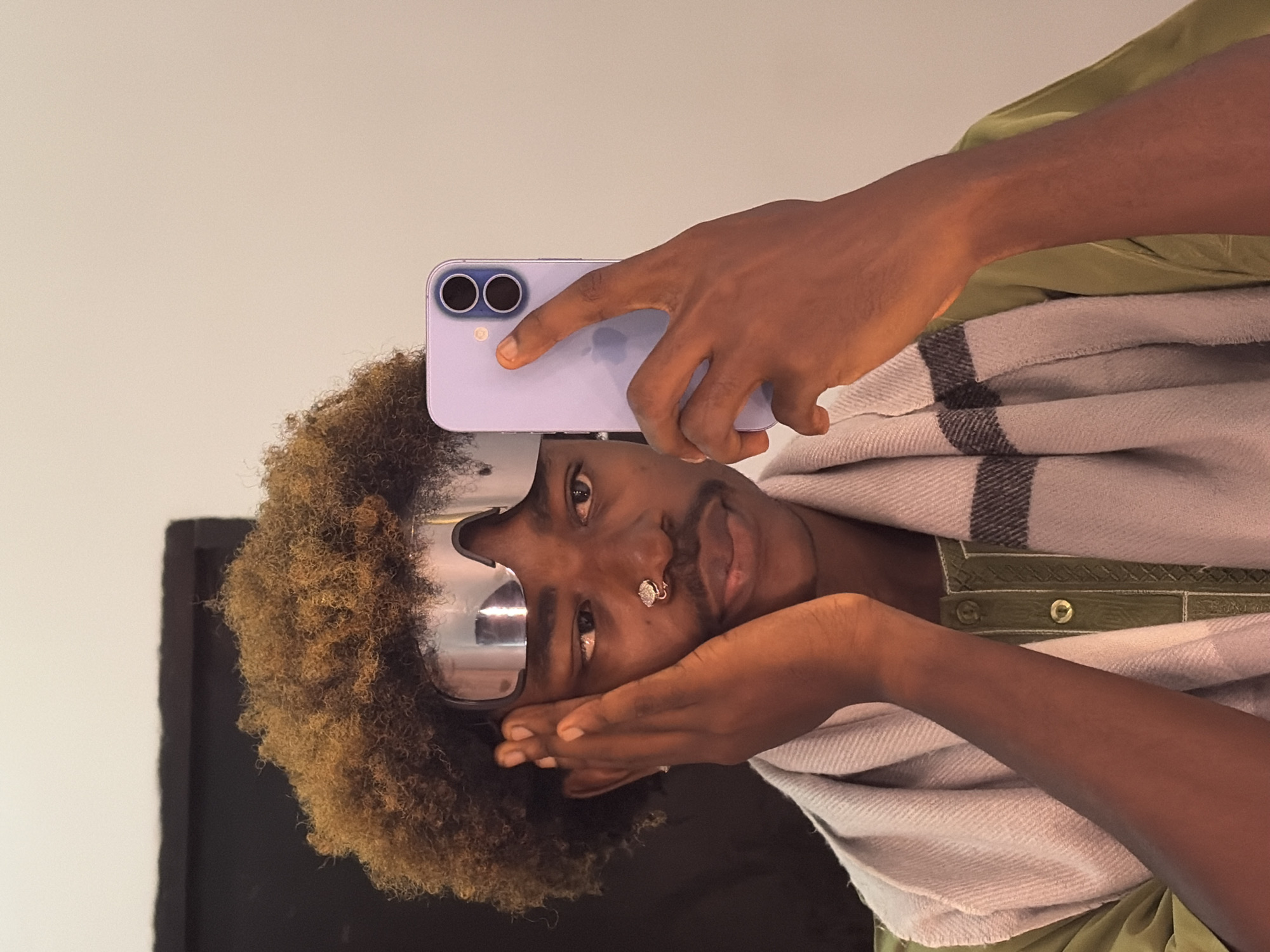An individual with dark skin and an afro-textured hairstyle with light brown highlights is depicted taking a mirror selfie indoors. They wear reflective silver visor-style sunglasses pushed onto their forehead, a double-hoop nose ring on their left nostril, and a small earring in their left ear. A thin mustache is present. The individual is dressed in a light olive green shirt with a patterned placket and a light grey scarf with dark grey striped accents draped around their neck. A purple iPhone, displaying two camera lenses on its rear, is held vertically in the individual's left hand to capture the image. Their right hand rests against their right cheek, fingers slightly spread. The individual's gaze is directed slightly downwards and to their left. The background consists of a plain, light-colored wall, with a dark, rectangular object partially visible in the upper left quadrant. The scene occurs indoors.
