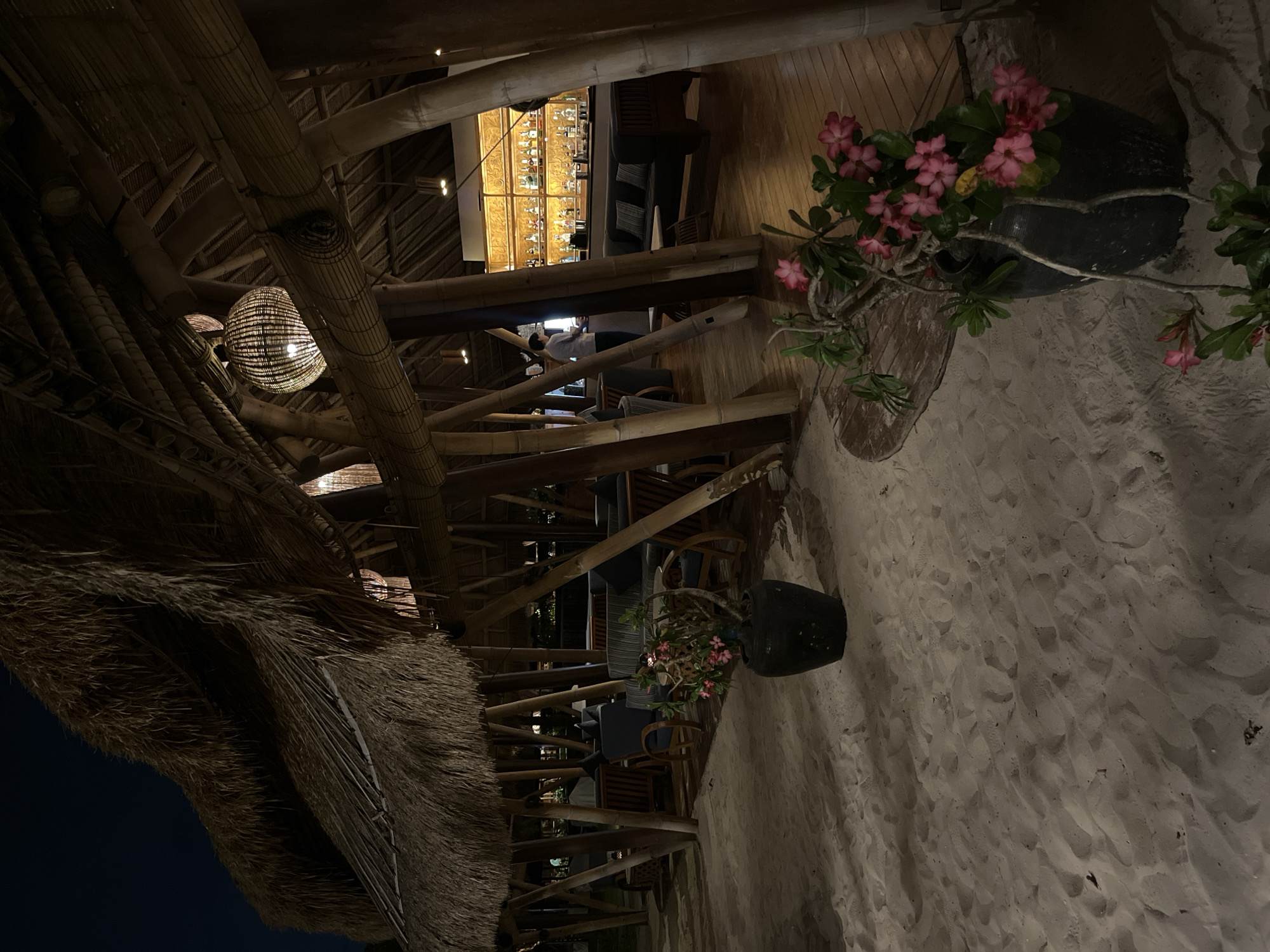 The image depicts a dimly lit, open-air establishment with a thatched roof supported by bamboo poles, likely located on a beach in Kuta Selatan, Indonesia. The foreground shows a sandy area with two large black planters containing flowering plants with pink blossoms. Beyond the sand, a wooden deck leads into the interior of the structure.
Inside, a bar area is visible with a well-stocked shelf of bottles illuminated by warm, golden lights. A person, dressed in a light-colored shirt and dark pants, stands behind the bar, appearing to be engaged in work. Comfortable seating, including sofas and chairs with dark upholstery and wooden frames, are arranged in various sections of the establishment. Woven pendant lights hang from the ceiling, adding to the ambient lighting. The overall atmosphere suggests a relaxed, tropical evening setting.