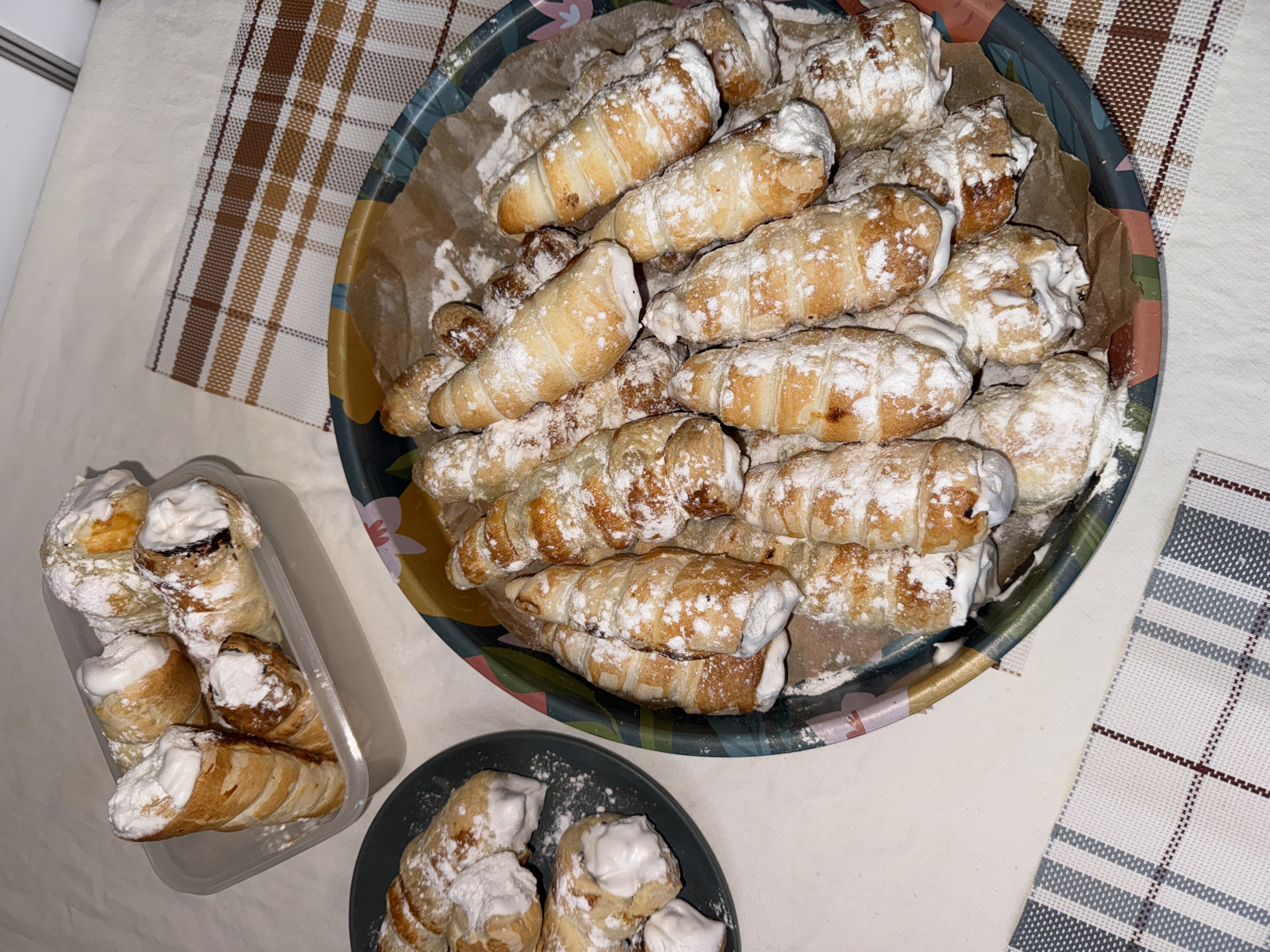 The scene, specified to be in Krasnoyarsk, Russia, depicts an indoor setting featuring multiple conical pastries, identified as cream horns, generously dusted with powdered sugar. In the foreground, a round tray with a dark rim and an interior multicolored floral or abstract pattern holds a large quantity of these pastries, resting on a sheet of light brown parchment paper. To the left of the tray, a smaller, dark grey round plate contains approximately five additional cream horns. In the upper left quadrant, a translucent rectangular plastic container holds several more pastries. All items are arranged on a light-colored surface, consistent with a table or countertop. Two distinct plaid placemats are partially visible: one with brown and white checks positioned in the upper right, and another with grey and white checks in the lower left. No individuals are present in the image. The lighting indicates an artificial indoor light source.