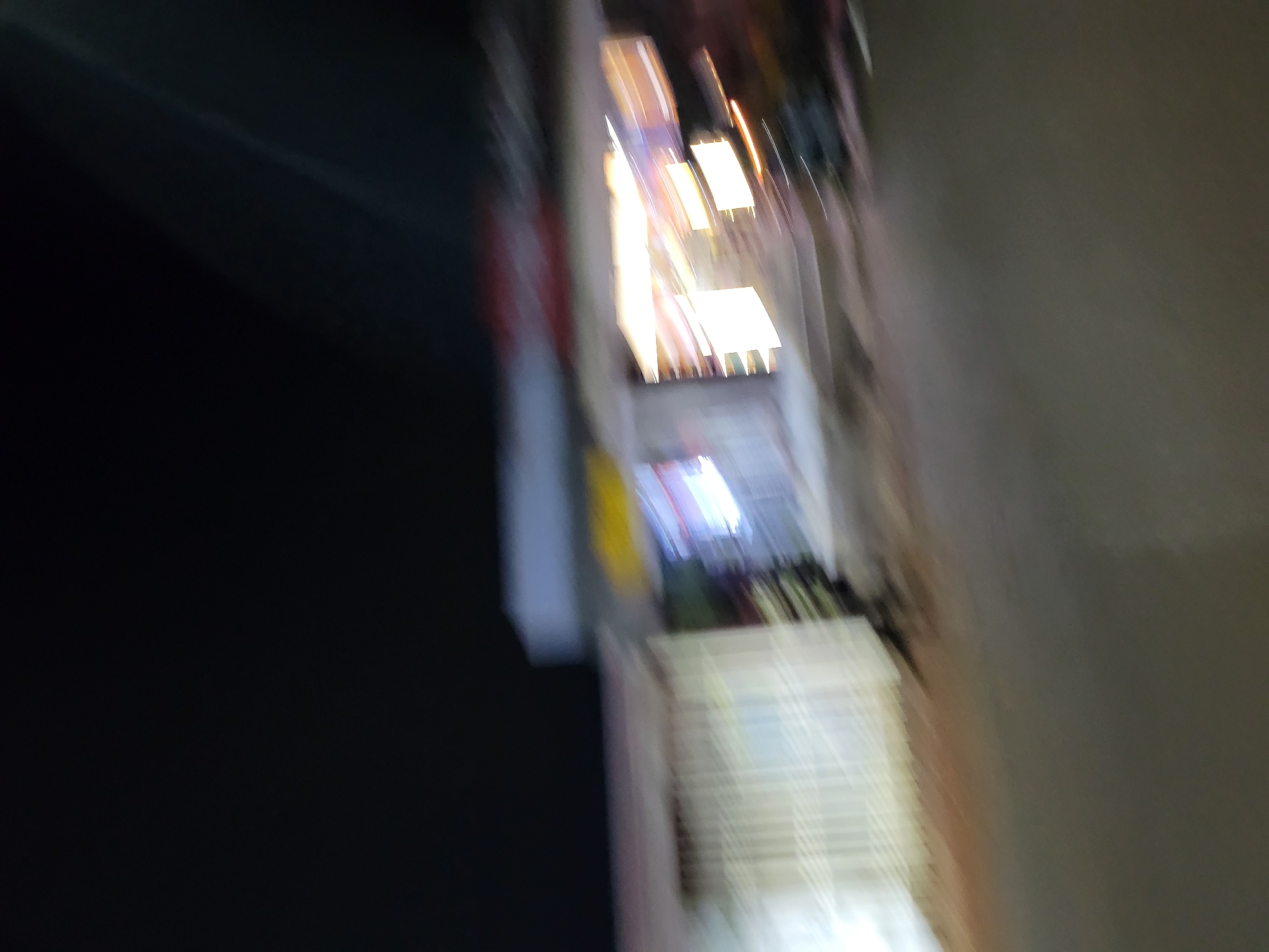 The image appears to be taken outdoors at night in an urban location. Several brightly lit storefronts line a street. There is dark asphalt in the foreground, likely a road or sidewalk. The rest of the environment is obscured by blur and poor lighting.