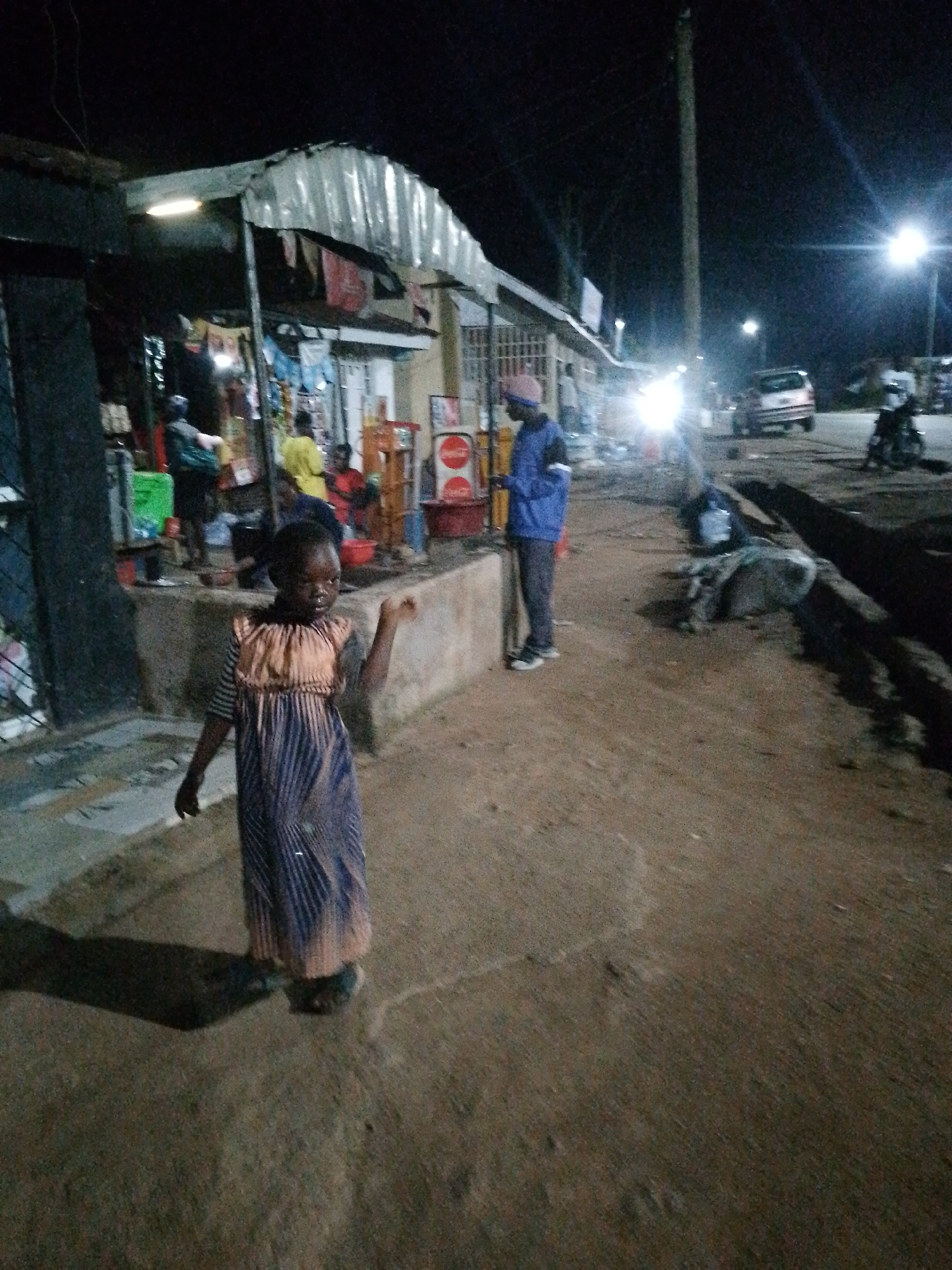 A young girl stands in the foreground of this nighttime image, looking towards the viewer with a slightly quizzical expression. She is wearing a patterned dress with a light-colored, pleated top and a striped skirt. To her left, a stall with a corrugated metal roof is visible, dimly lit by a single overhead light. Merchandise is displayed on shelves, and several people are blurred in the background, seemingly engaged in shopping or selling. Further in the background, a street scene unfolds. A person in a blue jacket and pink hat stands near a red and white object, possibly a beverage dispenser. A car is parked in the middle ground, illuminated by bright headlights. A motorcycle is also visible on the right side of the frame. The ground appears to be dirt or gravel, and shadows are cast by the lighting, indicating it's nighttime. The overall atmosphere is that of a busy market or street-side vendor area after dark. There is some blurry text on a sign to the right of the image, but it is illegible.