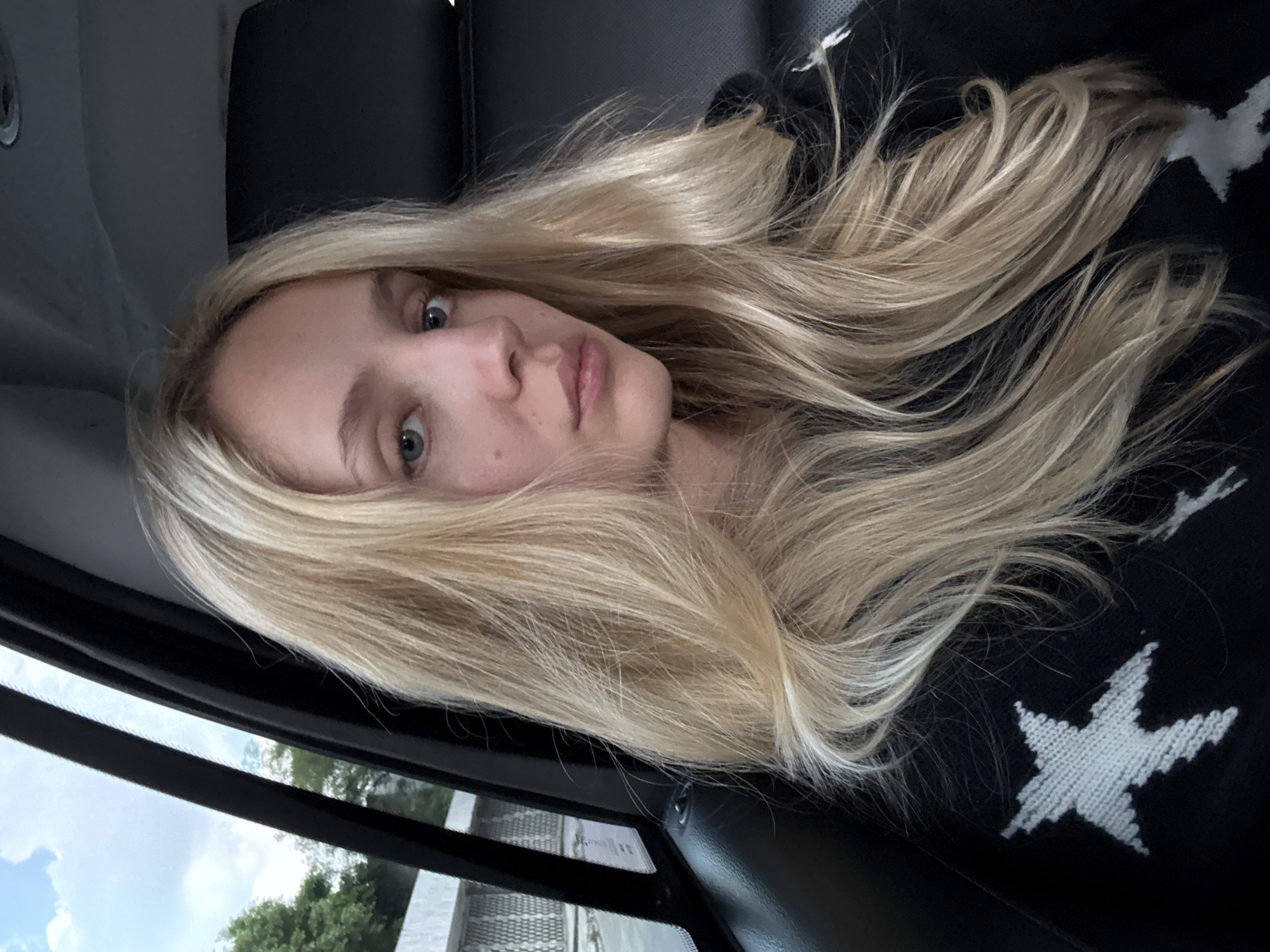 A woman with long blonde hair is sitting in the back seat of a vehicle. She is looking at the camera. The interior of the vehicle is dark leather. Outside the window, trees and a fence are visible. The sky is cloudy. The woman is wearing a black sweatshirt with white stars on it. She is not wearing makeup. The image appears to have been taken during the day.