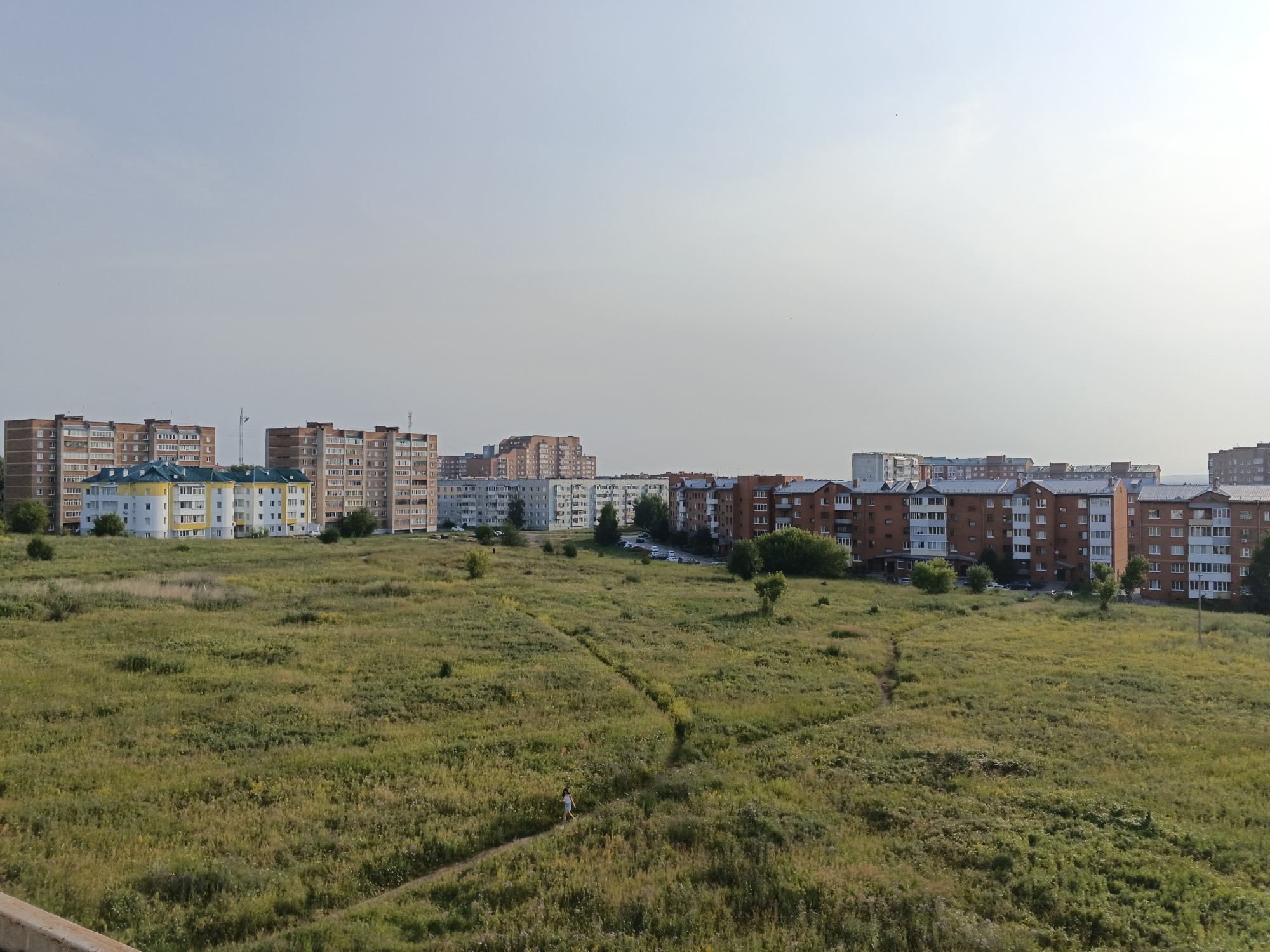 This is a photograph of a residential area in Yurga, Russia. The image features a large, open field of tall green grass and wildflowers in the foreground. A narrow, winding dirt path cuts through the field. A single person, wearing a white top and dark shorts, is walking on the path. In the background, a row of multi-story apartment buildings lines the horizon. The buildings are varied in color and architectural style, with some appearing to be made of brick and others of lighter colored materials. Trees are scattered throughout the landscape, and a few cars are visible on a road behind the apartment buildings. The sky is a hazy, pale blue. The scene suggests a peaceful, everyday moment in a suburban or semi-urban environment.