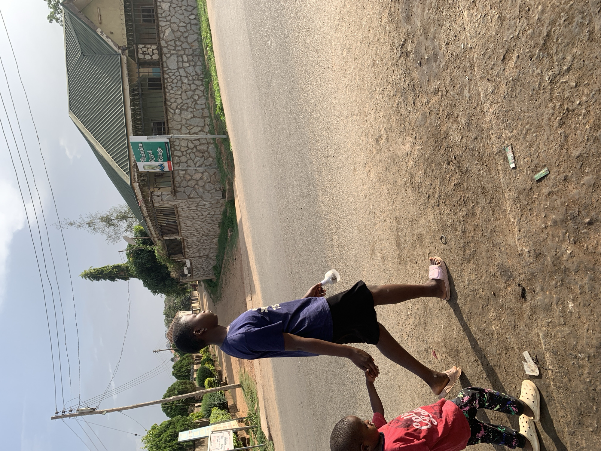 Two children are walking hand-in-hand on a road in Gura Top, Nigeria. The taller child, wearing a blue shirt and black shorts, holds a white object in their other hand and wears pink slippers. The shorter child wears a pink shirt and patterned leggings, with light-colored clogs on their feet. They are walking on a paved road with a rough shoulder. A building made of stone with a green roof and a sign that reads "Christian Royal College" is visible. There are utility poles and wires, trees, and other buildings in the background. Debris is on the road.
