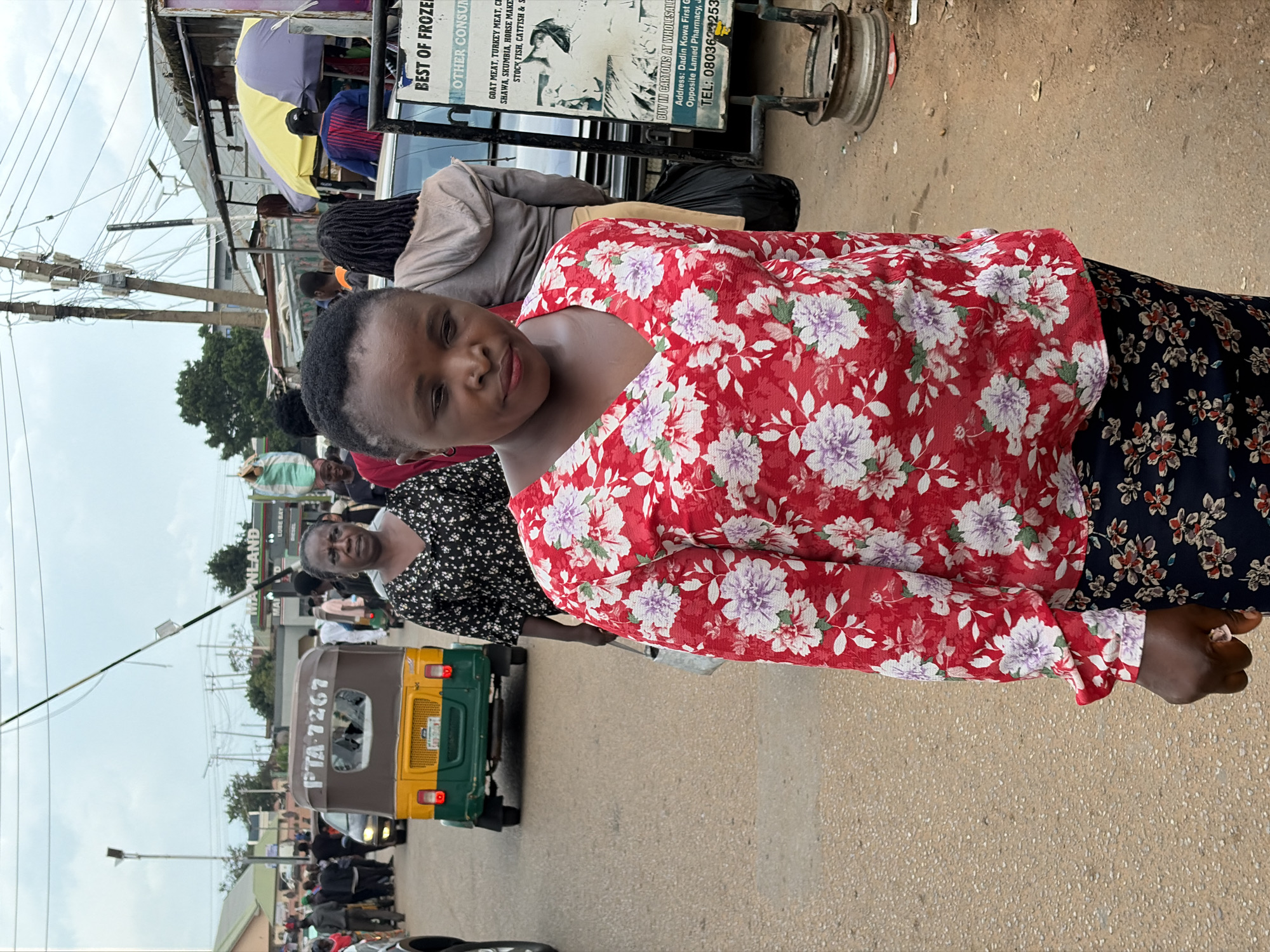 A medium shot captures a bustling street scene in Jos, Nigeria, with a focus on several individuals. In the foreground, a woman wearing a red floral blouse and a dark floral skirt walks towards the viewer, her gaze slightly downward. To her left and slightly behind her, another woman in a black and white floral top is visible. Behind them, a yellow and green auto-rickshaw with the license plate "PTA 7267" is in motion. The background reveals more people walking and standing, a variety of stalls and shops, a gas station sign that reads "MAINLAND" and "LUBE BAY", and overhead electrical wires. A sign board in the lower right corner advertises "BEST OF FROZEN OTHER CONSUM". The ground is paved with asphalt and gravel. The sky is overcast, suggesting a cloudy day. The overall impression is one of everyday urban activity in a Nigerian city.